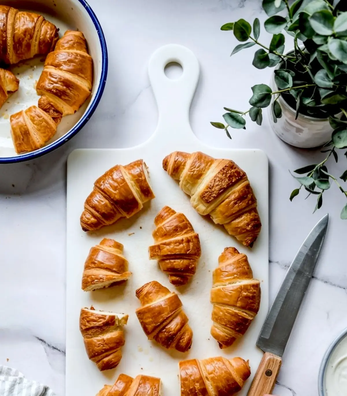 Golden brown croissants arranged on a white cutting board with a kitchen knife and a potted plant nearby, creating a cozy breakfast setting.