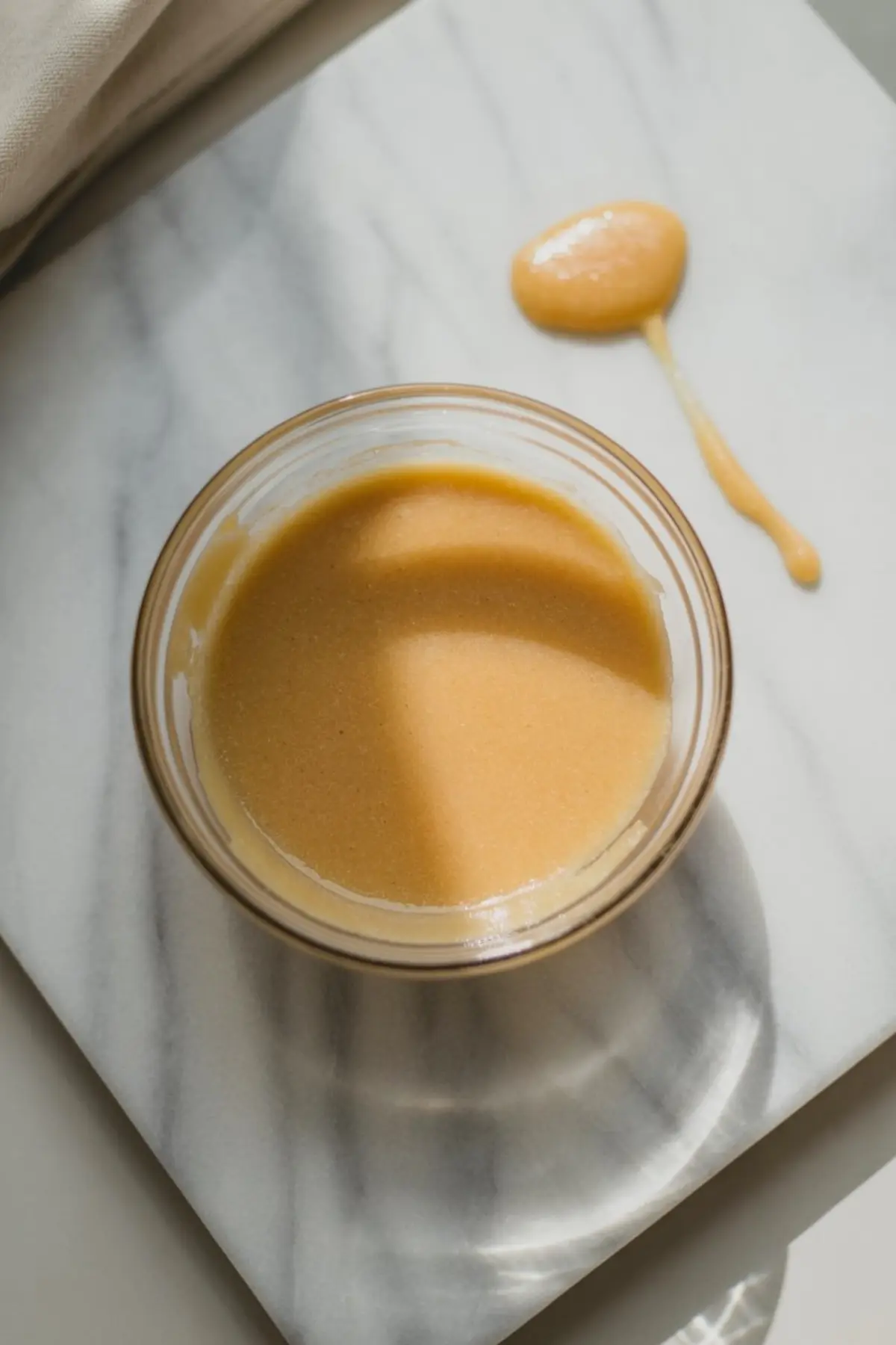 Small glass bowl of smooth maple sugar mixture placed on a marble surface, with a spoonful of the mixture drizzled nearby.
