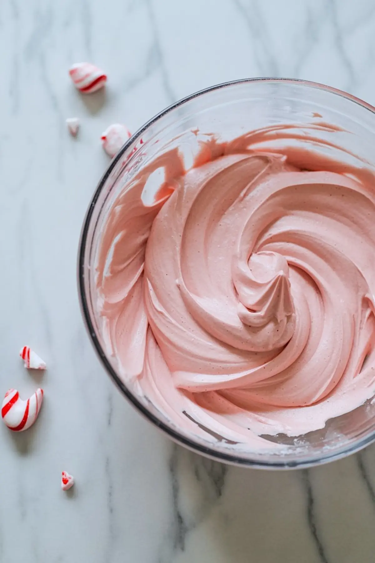 Smooth pink peppermint frosting swirled in a glass bowl on a marble surface, with crushed red and white candy cane pieces scattered nearby.
