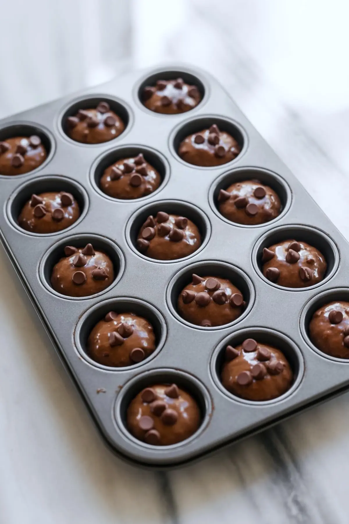 Muffin tin filled with chocolate brownie batter topped with chocolate chips, ready for baking on a marble counter.
