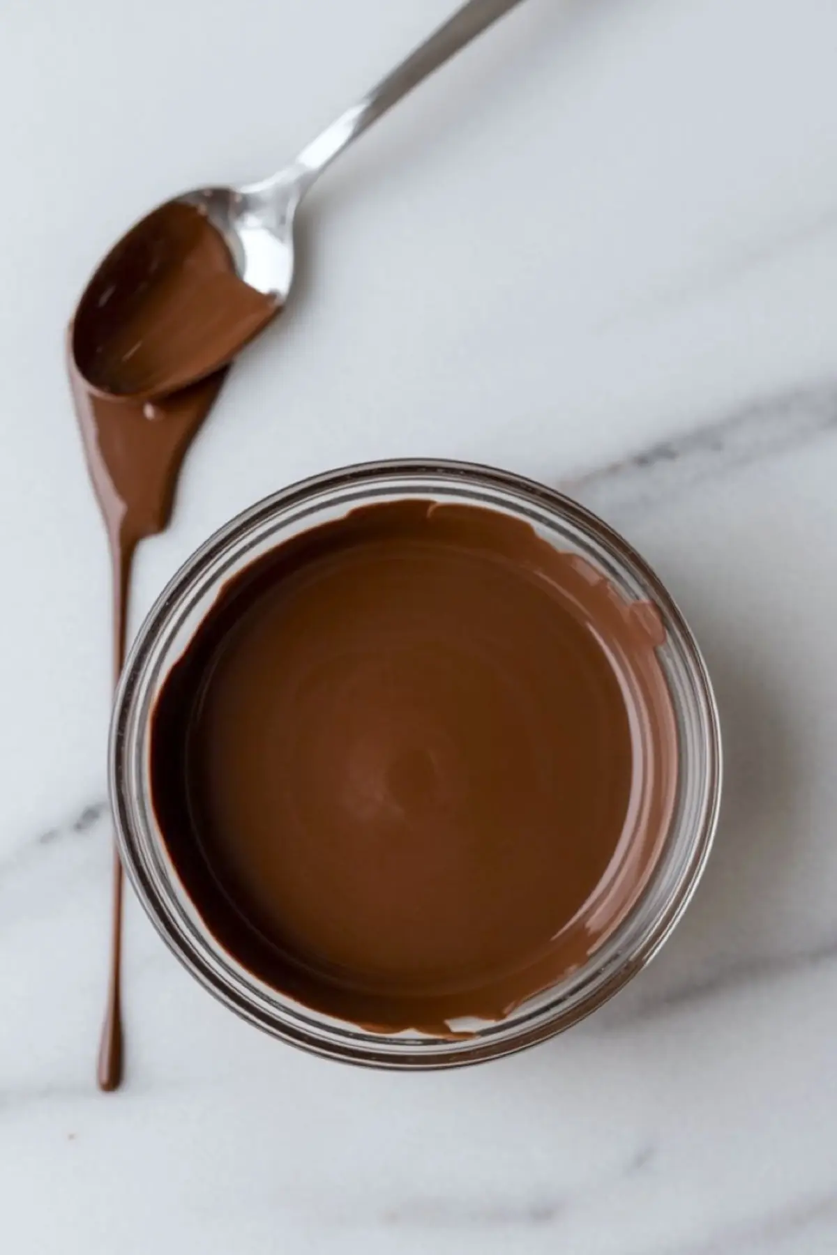 Small glass bowl filled with glossy melted chocolate, with a metal spoon resting beside it on a marble surface, showing a drizzle of chocolate.