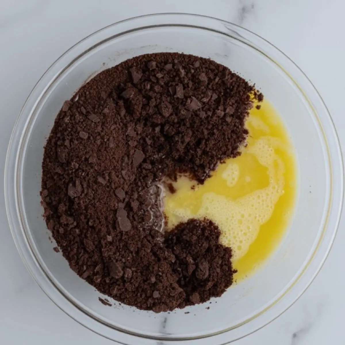 Overhead view of crushed chocolate cookie crumbs and melted butter partially combined in a clear glass bowl on a white countertop.