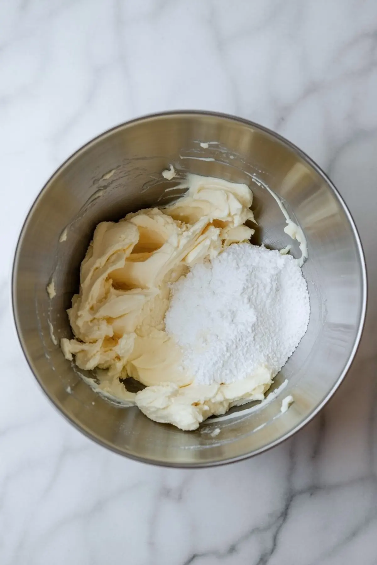 Mixing bowl filled with softened butter and powdered sugar on a marble countertop, ready for creaming.