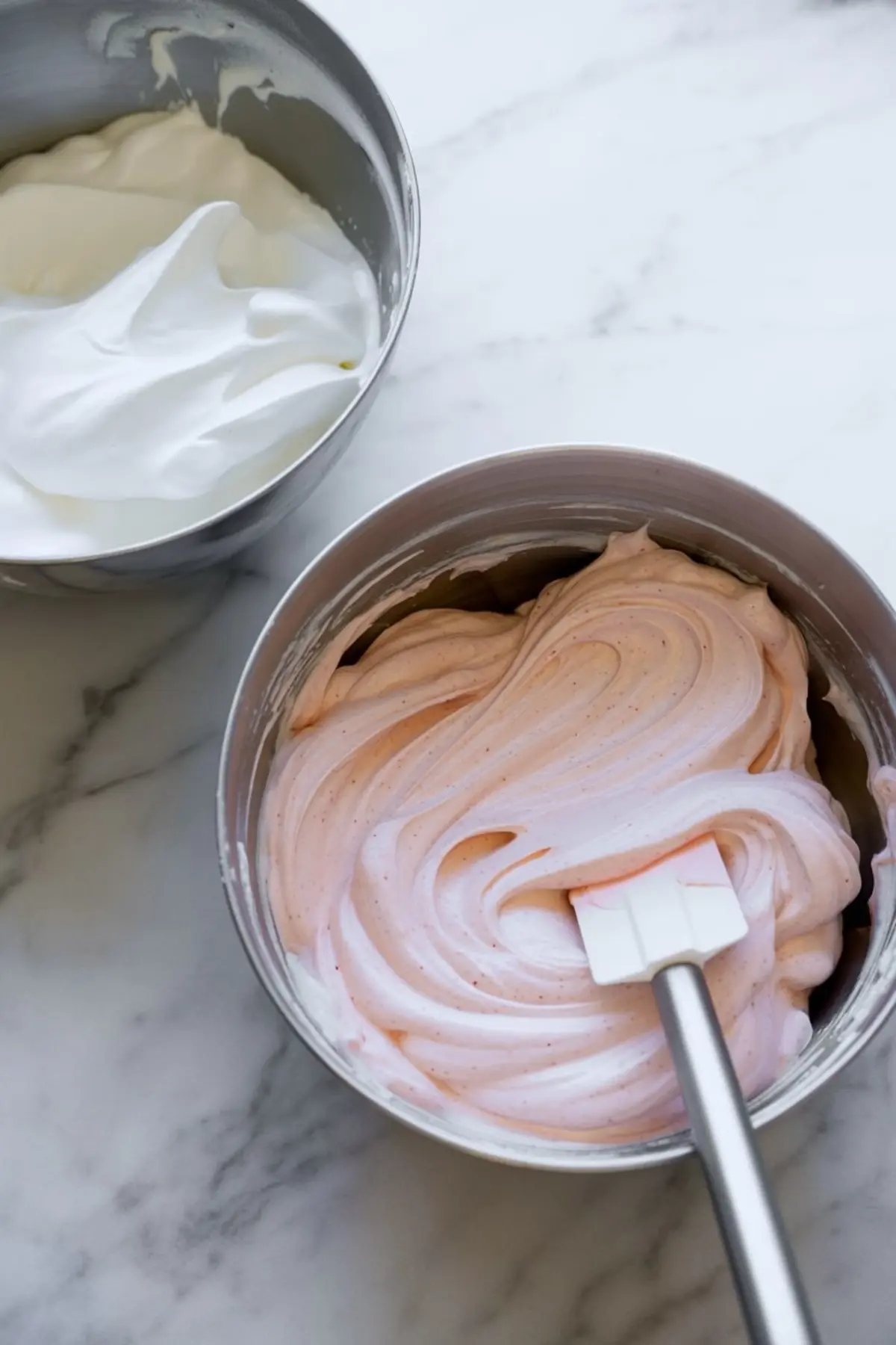 Two metal bowls on a marble surface, one with fluffy white whipped cream and the other with light pink peppermint filling being mixed with a spatula.