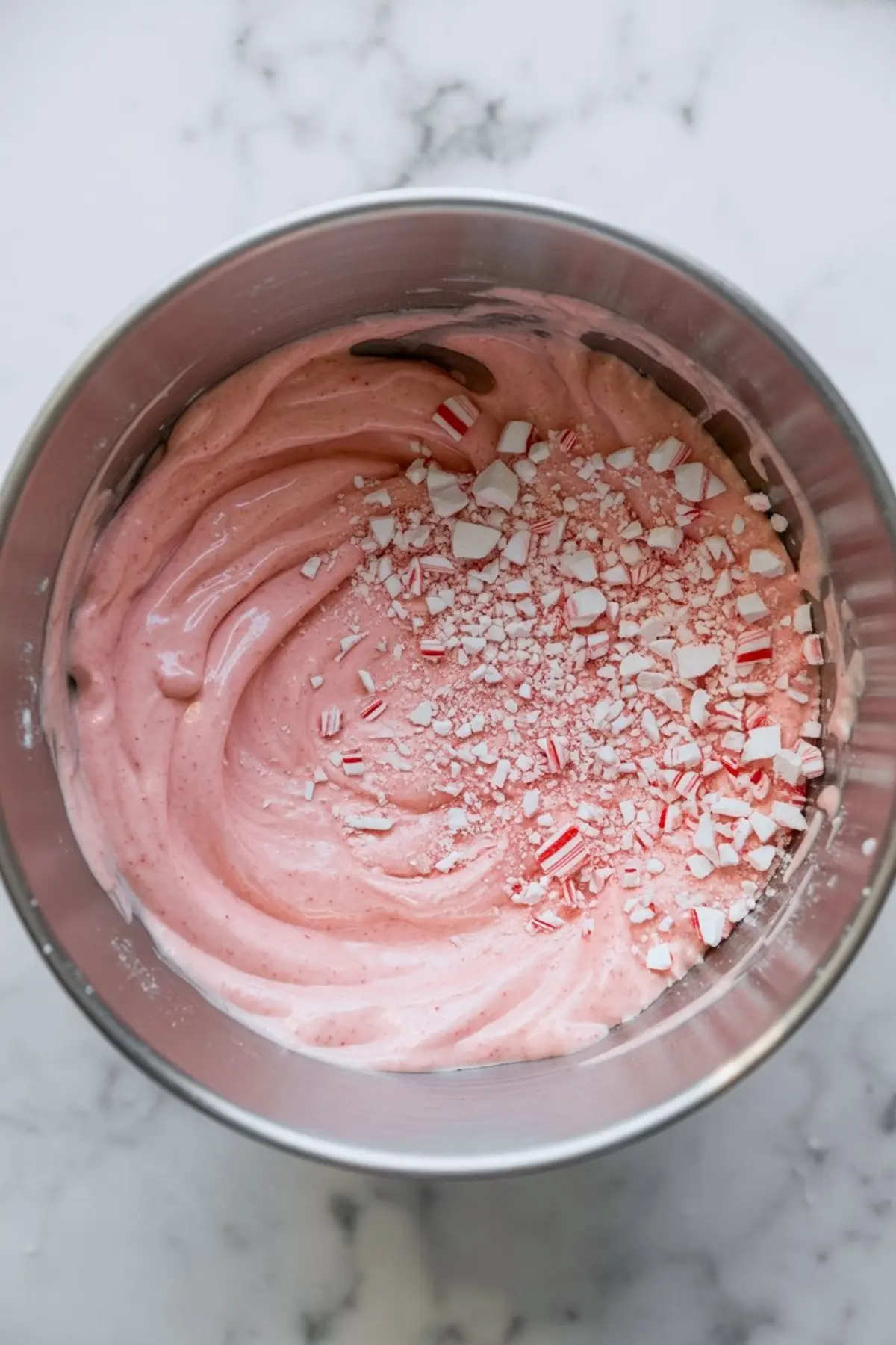 Crushed peppermint candies sprinkled over pink peppermint filling in a metal mixing bowl on a marble background.