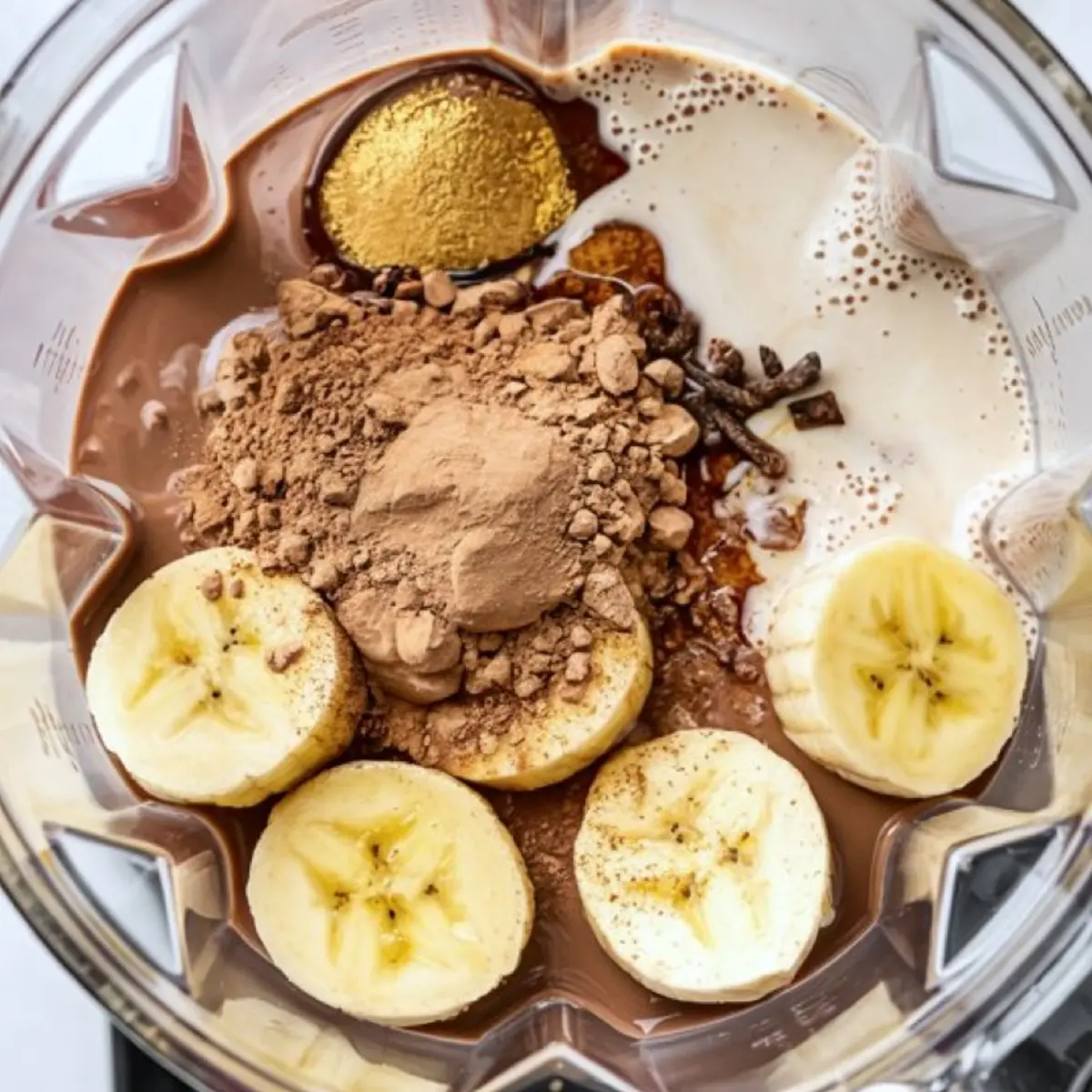 Overhead view of a blender filled with banana slices, cocoa powder, almond milk, maple syrup, cardamom pods, and chai spices for a chocolate chai smoothie.
