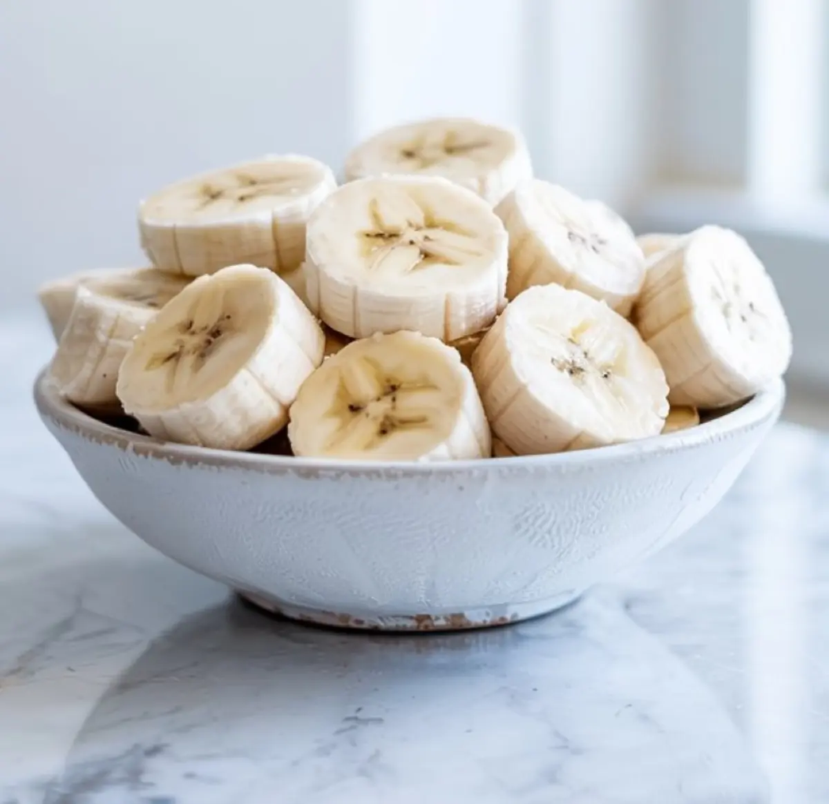 White ceramic bowl filled with fresh banana slices stacked on top of each other, placed on a glossy marble surface with soft natural lighting.
