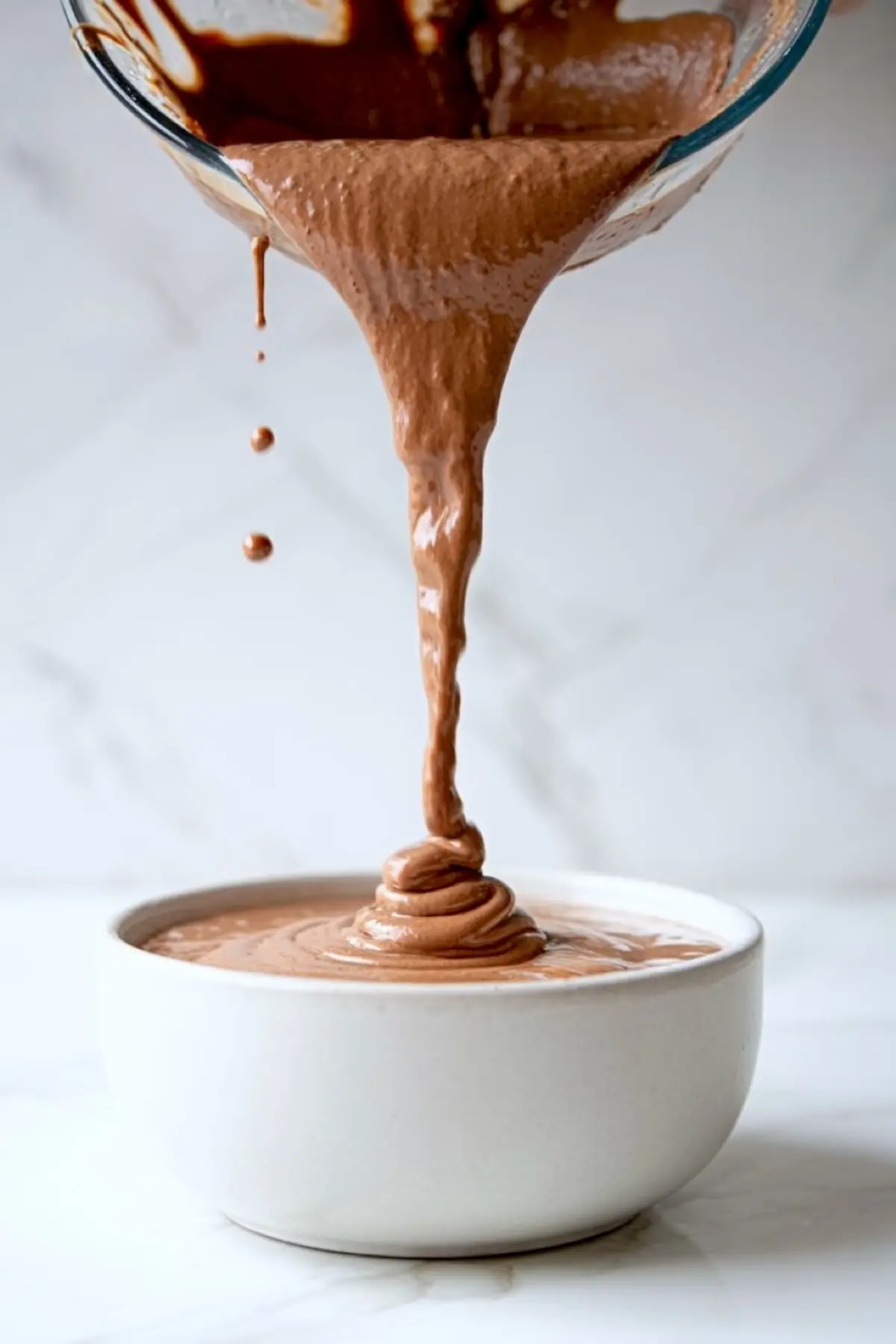 Thick chocolate chai smoothie being poured from a blender into a white bowl, creating a creamy swirl against a marble background.
