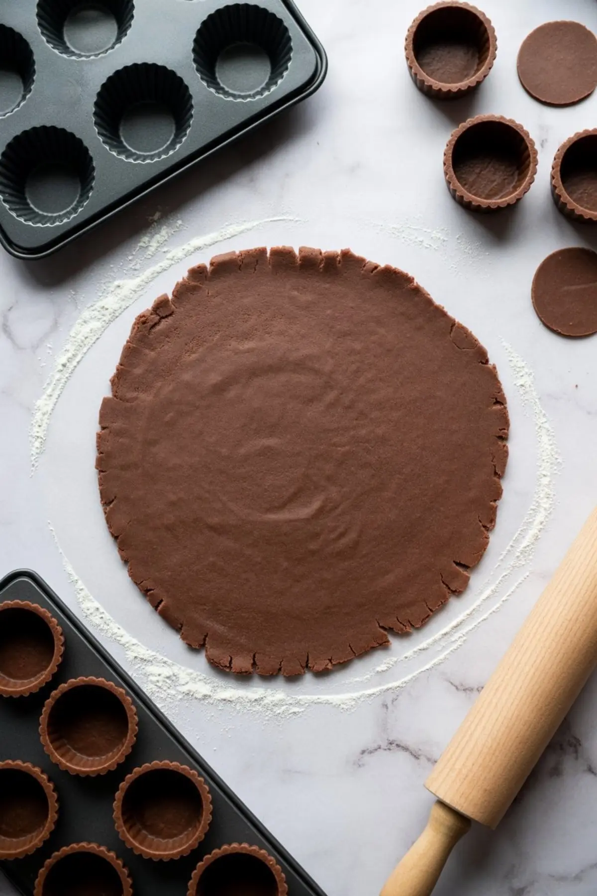 Rolled chocolate dough on a floured surface with cut tartlet shells and a rolling pin, surrounded by empty tart pans.
