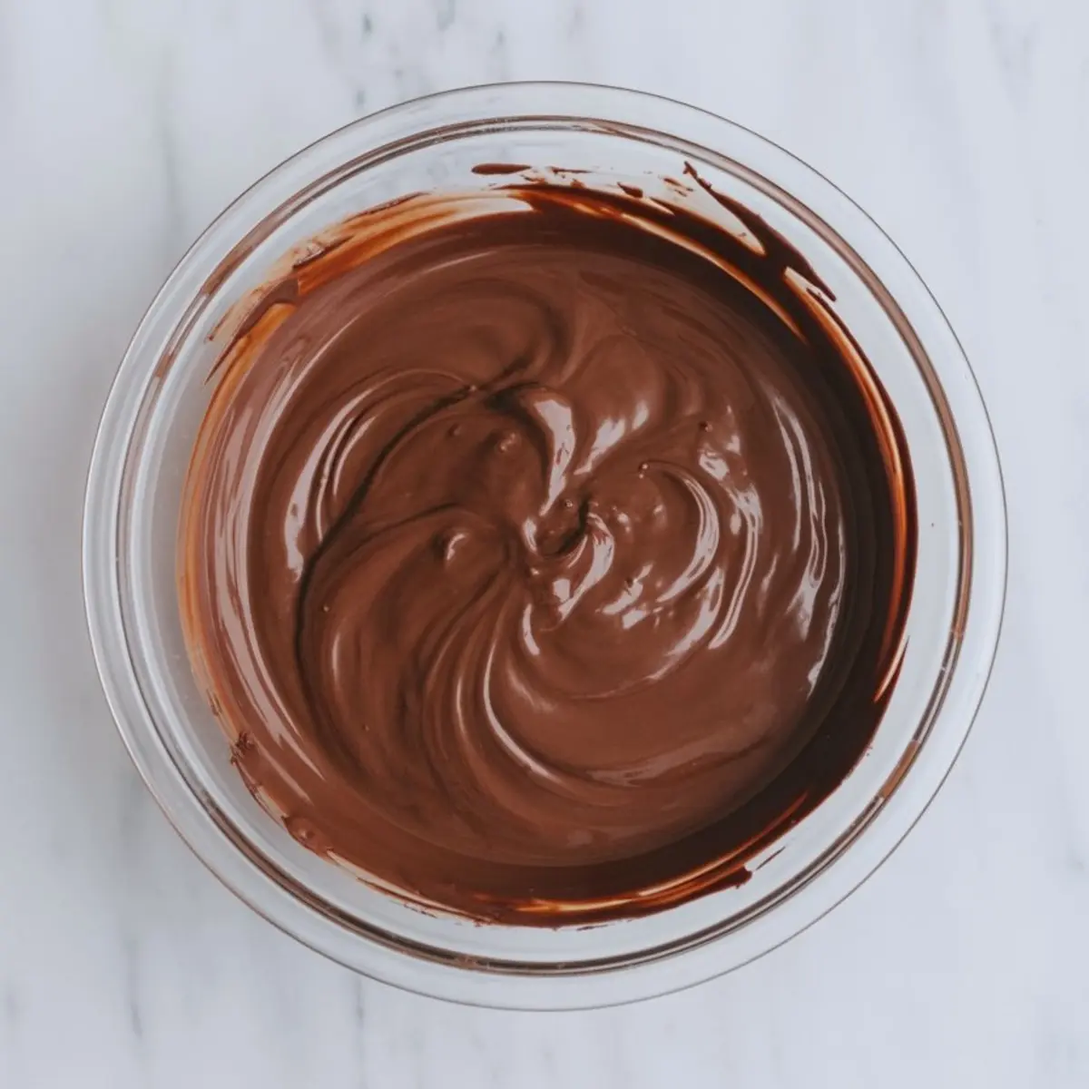 Smooth melted chocolate swirled in a glass bowl on a marble surface, ideal for baking ganache or dessert fillings.
