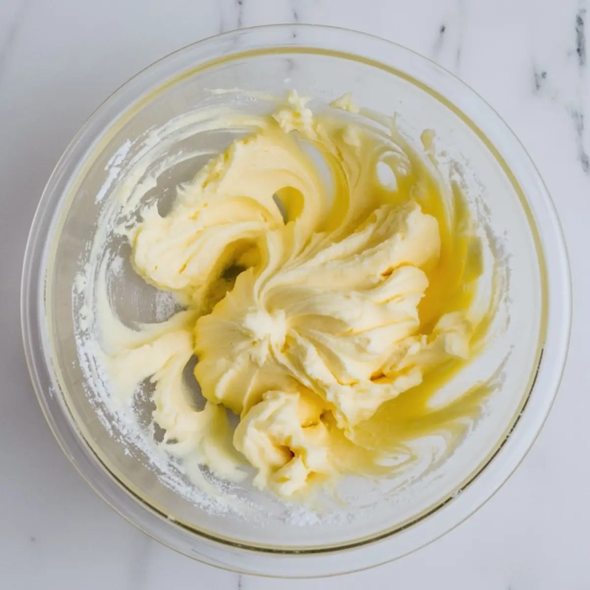 Creamed butter and sugar mixture swirled in a glass bowl, set on a white marble countertop, showing the beginning stage of cookie dough preparation.
