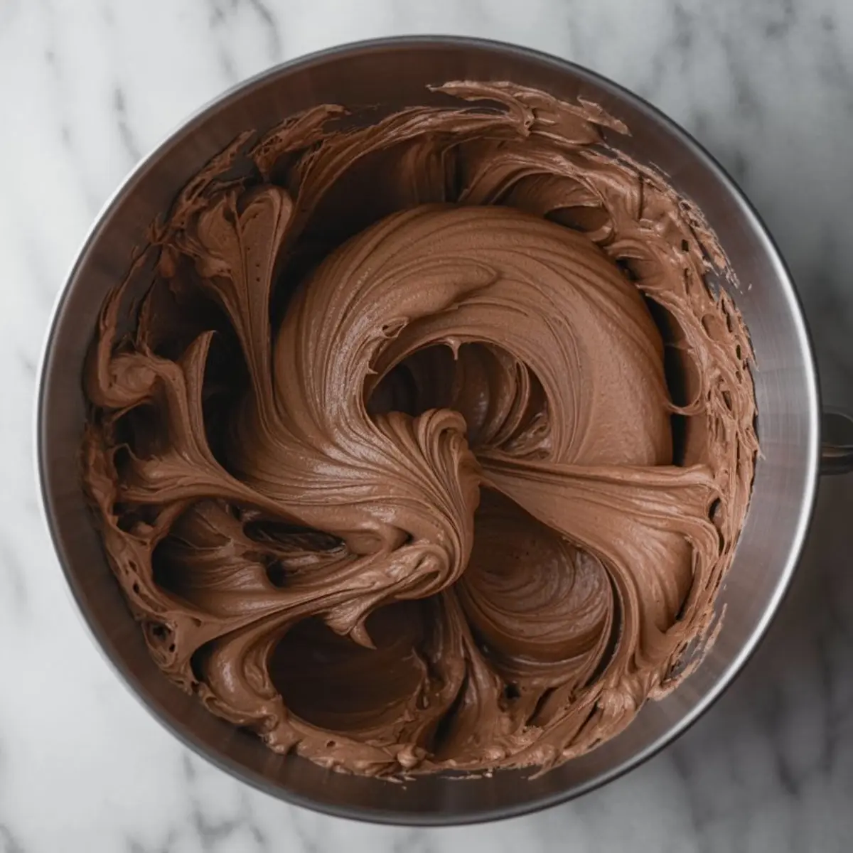 Thick chocolate fudge frosting swirled in a mixing bowl with a smooth, glossy texture, ideal for cupcakes or layered cakes.