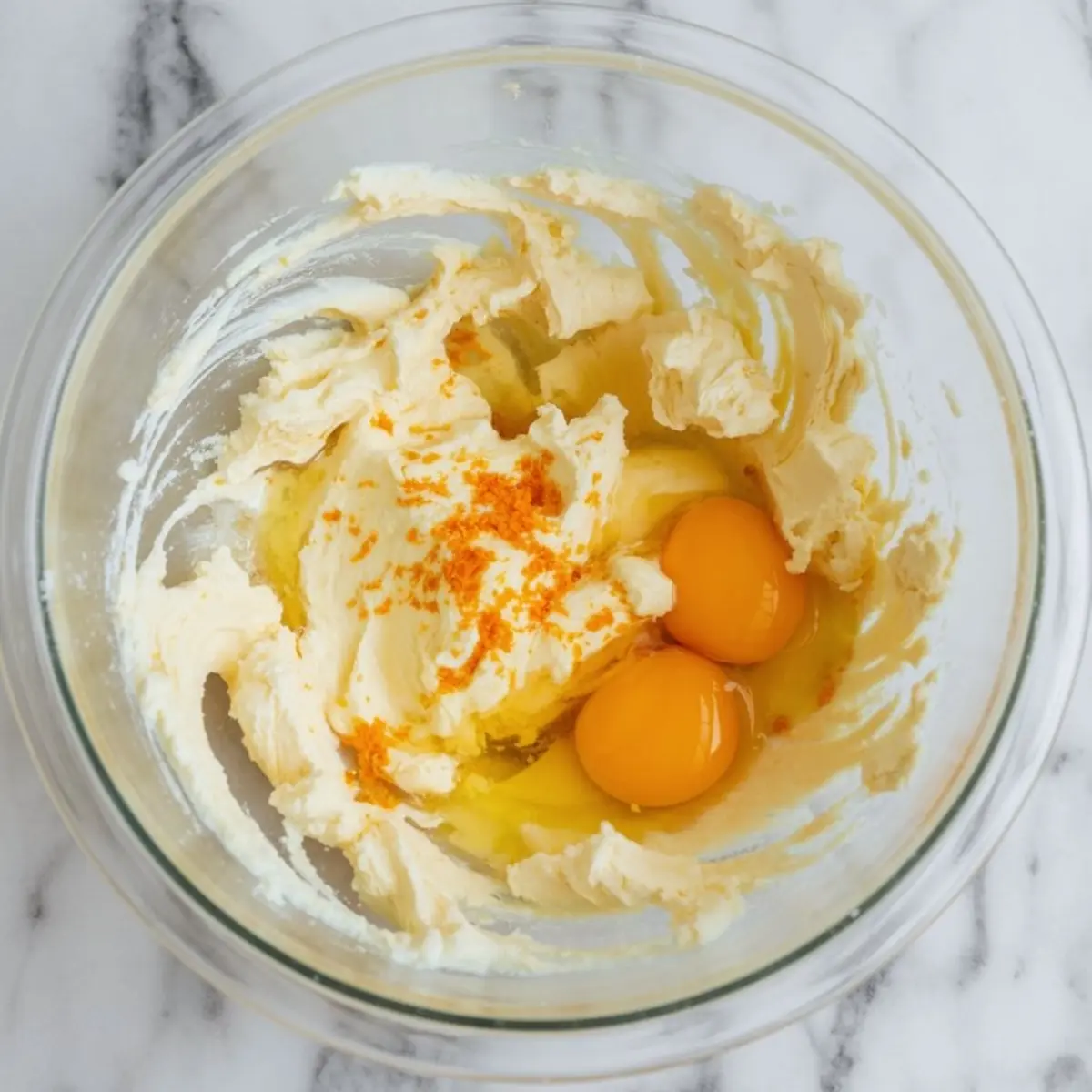 Glass bowl with butter and sugar creamed together, two cracked eggs, and fresh orange zest on a marble surface, showing early stages of cake batter preparation.
