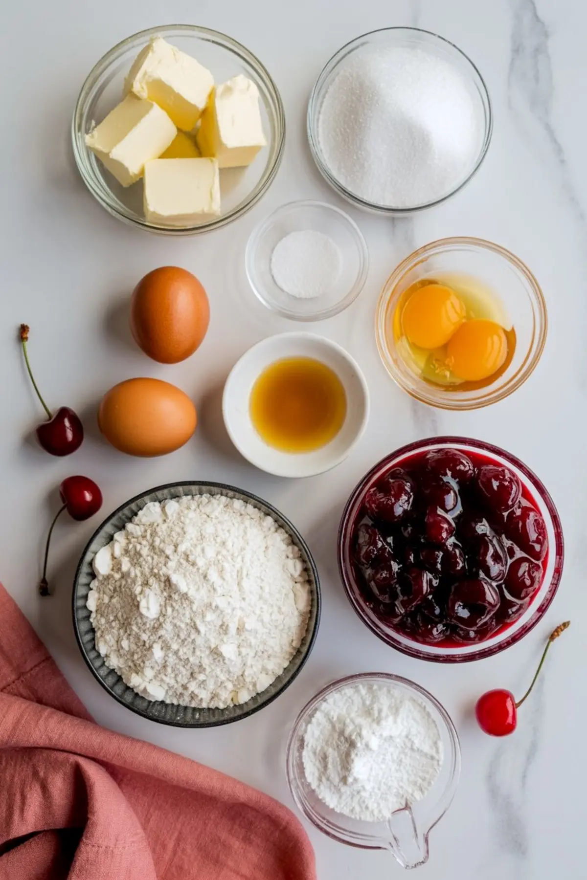 Flat lay of baking ingredients including flour, granulated sugar, powdered sugar, butter, eggs, vanilla extract, baking powder, and a bowl of cherry pie filling on a white marble surface.