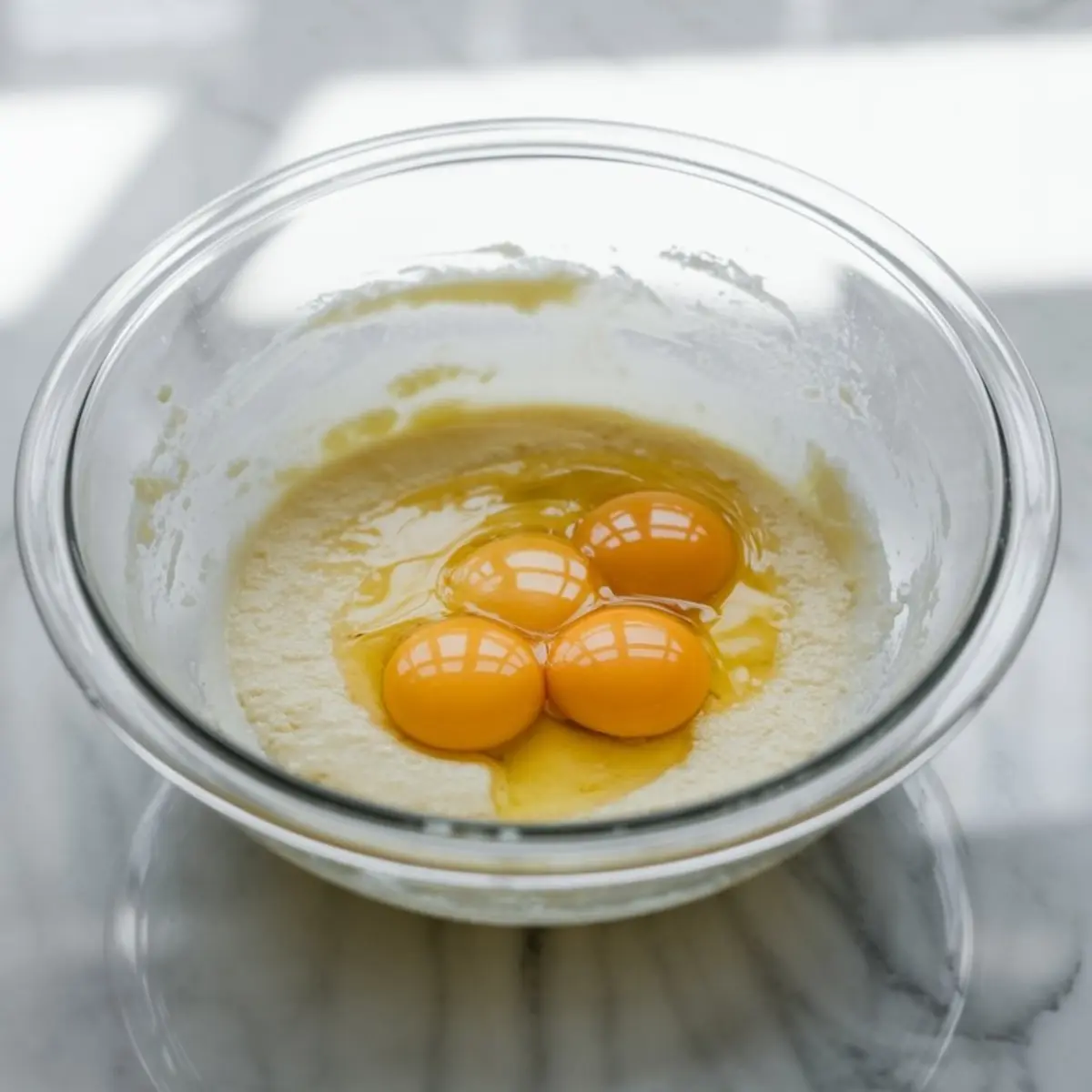 Four raw egg yolks sitting on top of a butter-sugar mixture in a glass bowl, part of a baking process for cakes or dessert bars.
