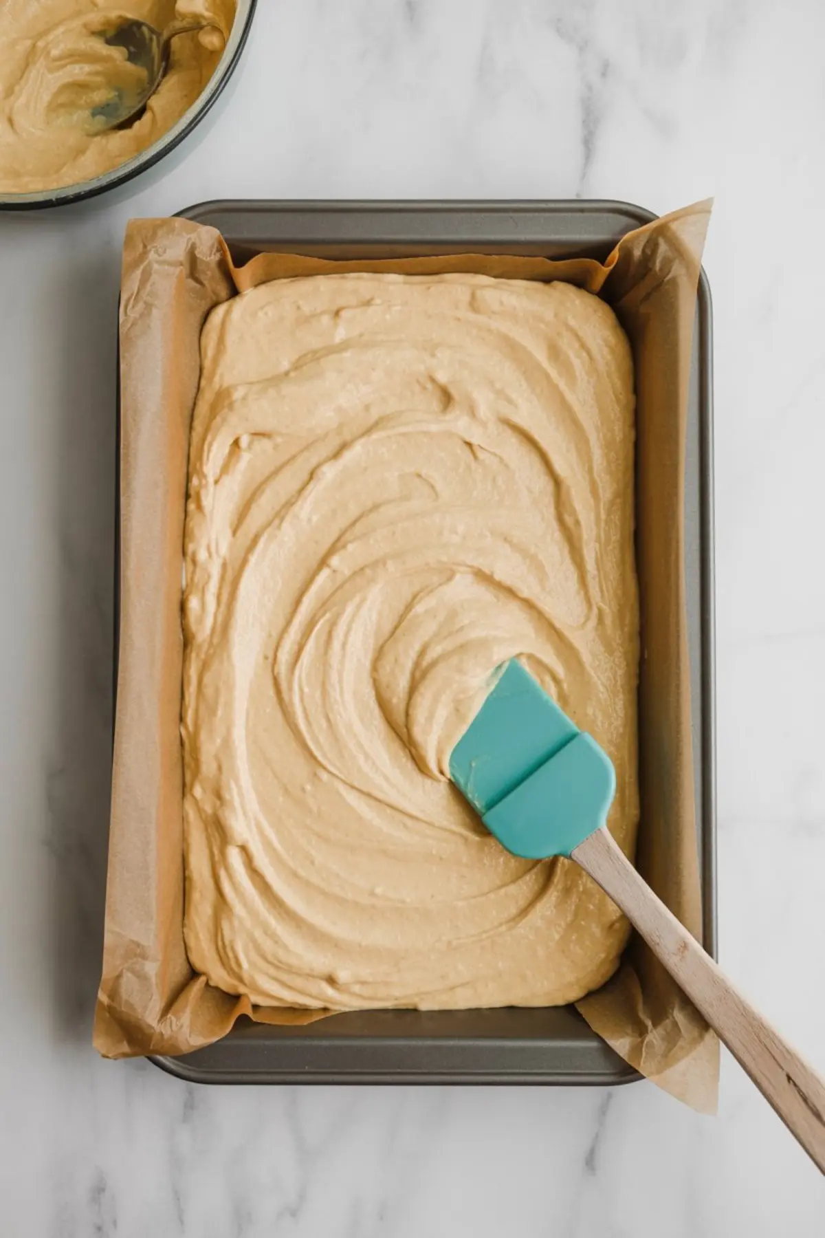 Cake batter spread evenly in a parchment-lined rectangular baking pan using a teal silicone spatula, ready for baking.