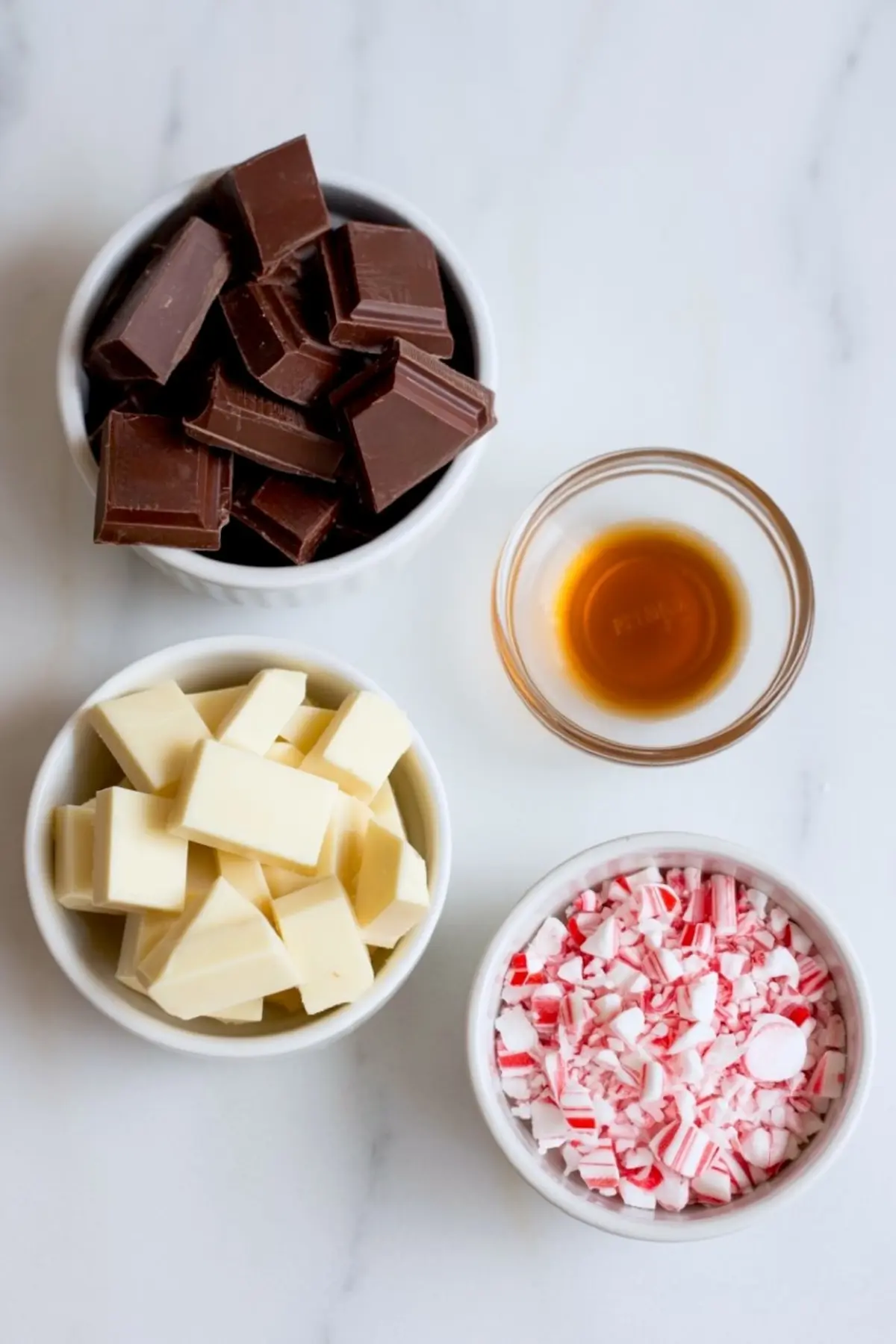 Top view of four small bowls with peppermint bark ingredients including milk chocolate chunks, white chocolate chunks, vanilla extract, and crushed candy canes on a white surface.
