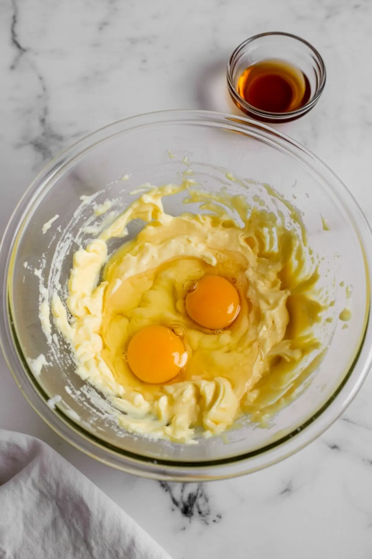 Glass mixing bowl with creamed butter and sugar mixture, cracked raw eggs on top, and a small bowl of vanilla extract placed nearby on a marble surface.