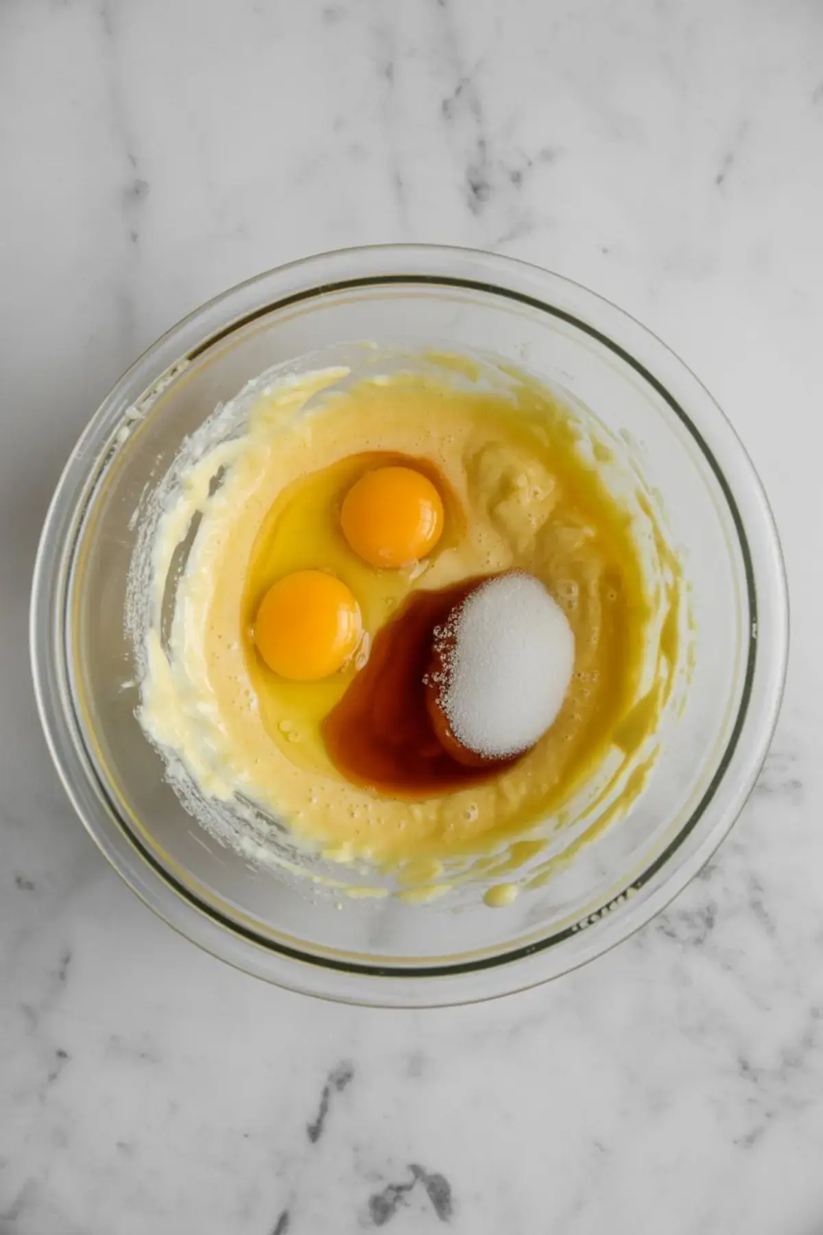 Glass mixing bowl with a yellow batter base, containing two whole eggs, sugar, and vanilla extract, ready to be blended for a cake recipe or cupcake batter. Ideal process shot for easy baking recipes or cake mixing tutorials.
