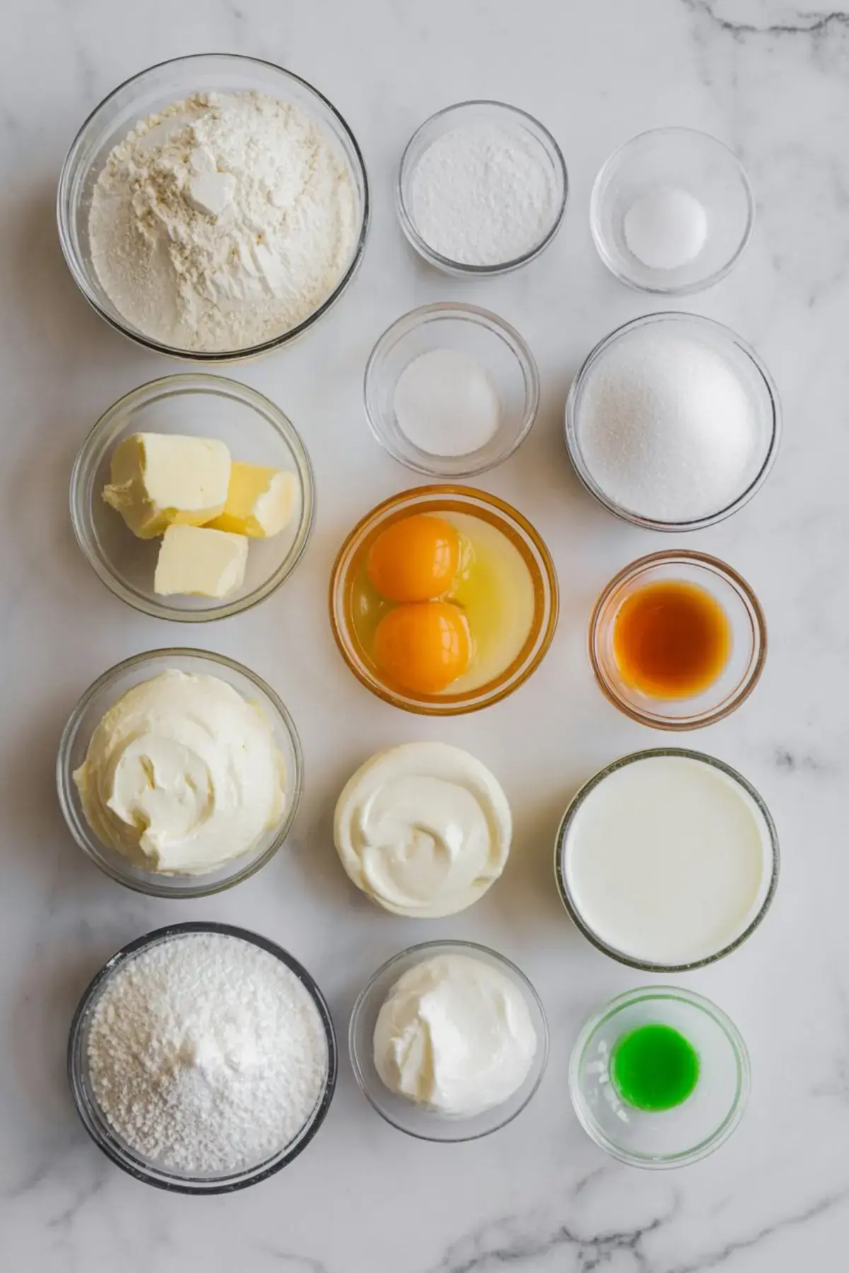 Flat lay of pre-measured baking ingredients in small glass bowls, including flour, sugar, butter, eggs, vanilla, sour cream, yogurt, and green food coloring. A complete ingredient lineup for frosting recipes, cupcakes, or holiday cakes.
