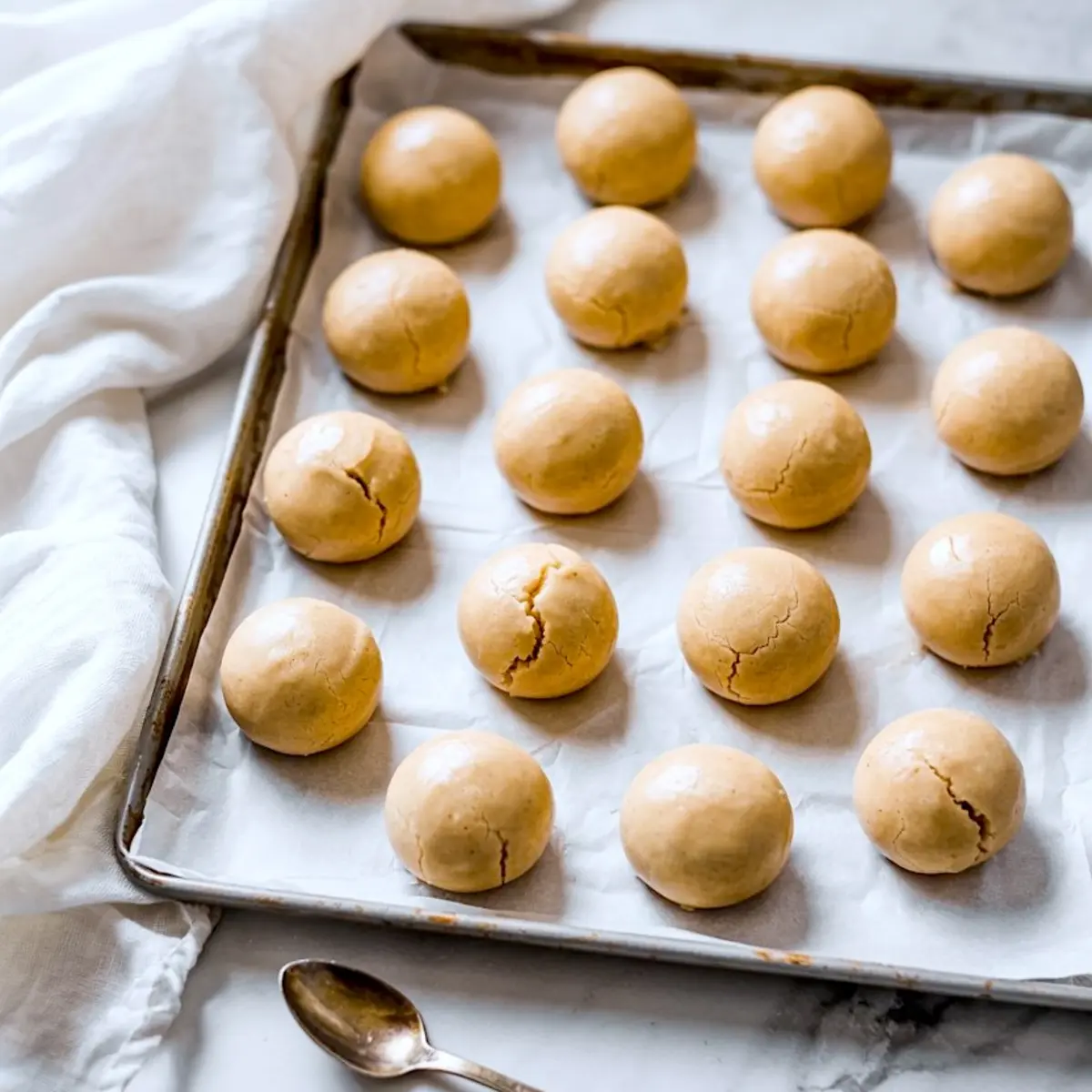 Baking tray lined with parchment paper displaying rows of unbaked round cookie dough balls with cracked tops.