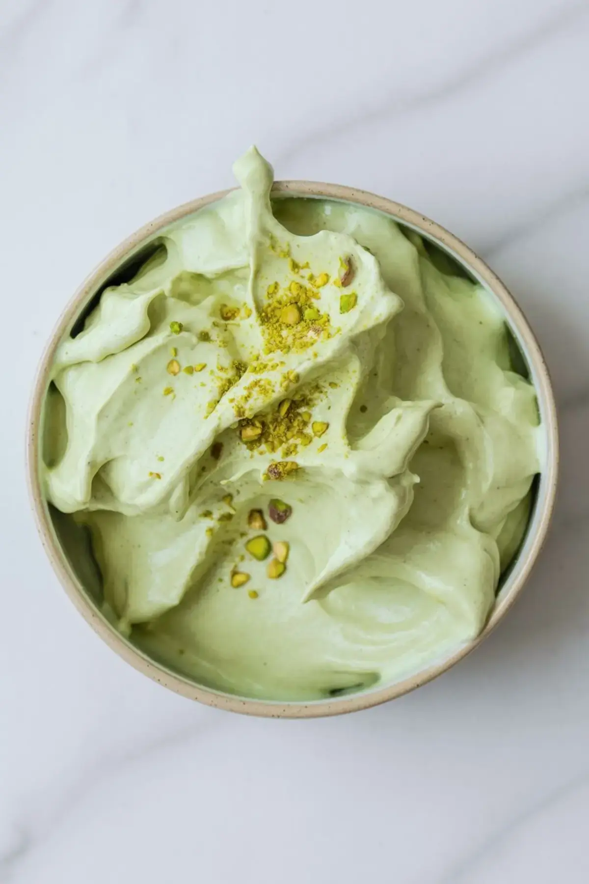 A ceramic bowl filled with smooth pistachio pastry cream shows a light green whipped texture topped with crushed pistachios, perfect for filling desserts like cream puffs or éclairs.
