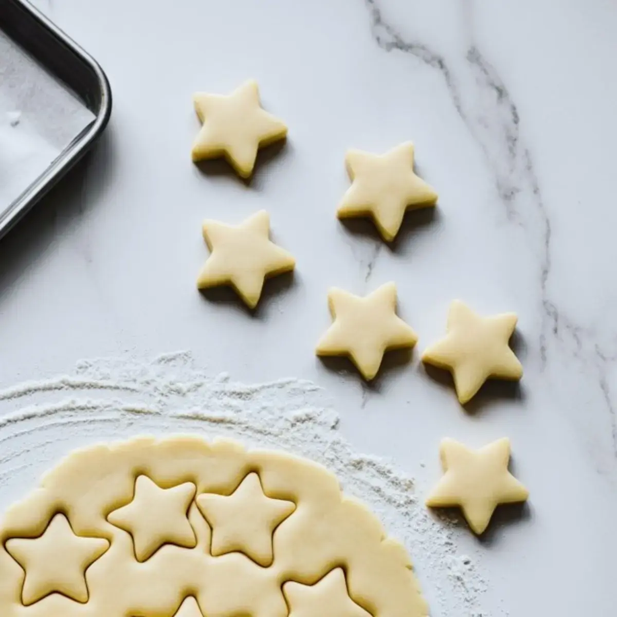 Star-shaped sugar cookie dough cutouts rest on a floured surface next to a metal baking tray, showing the preparation stage of cookie toppers for holiday-themed desserts.

