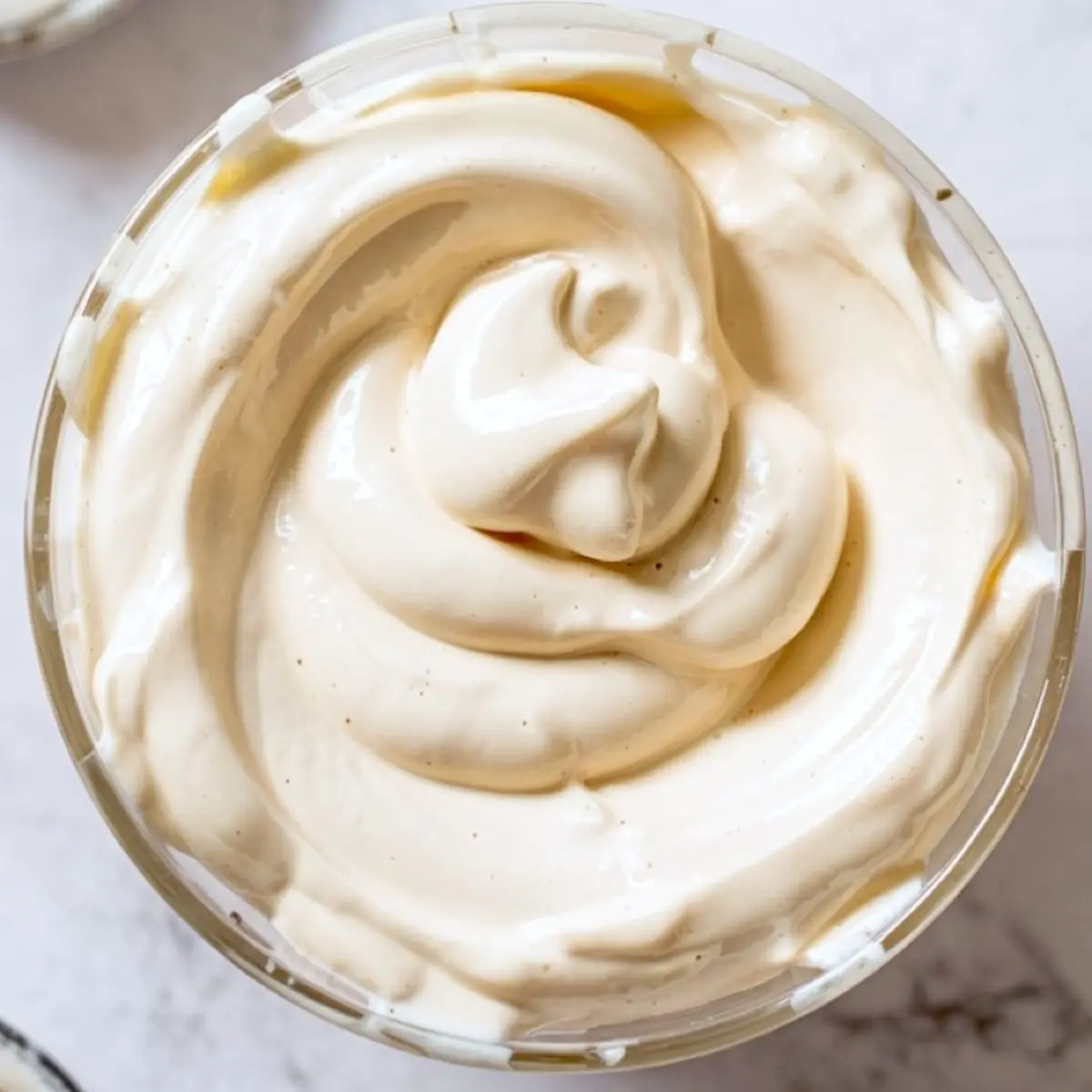 Overhead close-up of a glass bowl filled with thick, swirled whipped cream or mascarpone topping, with a smooth and creamy texture.