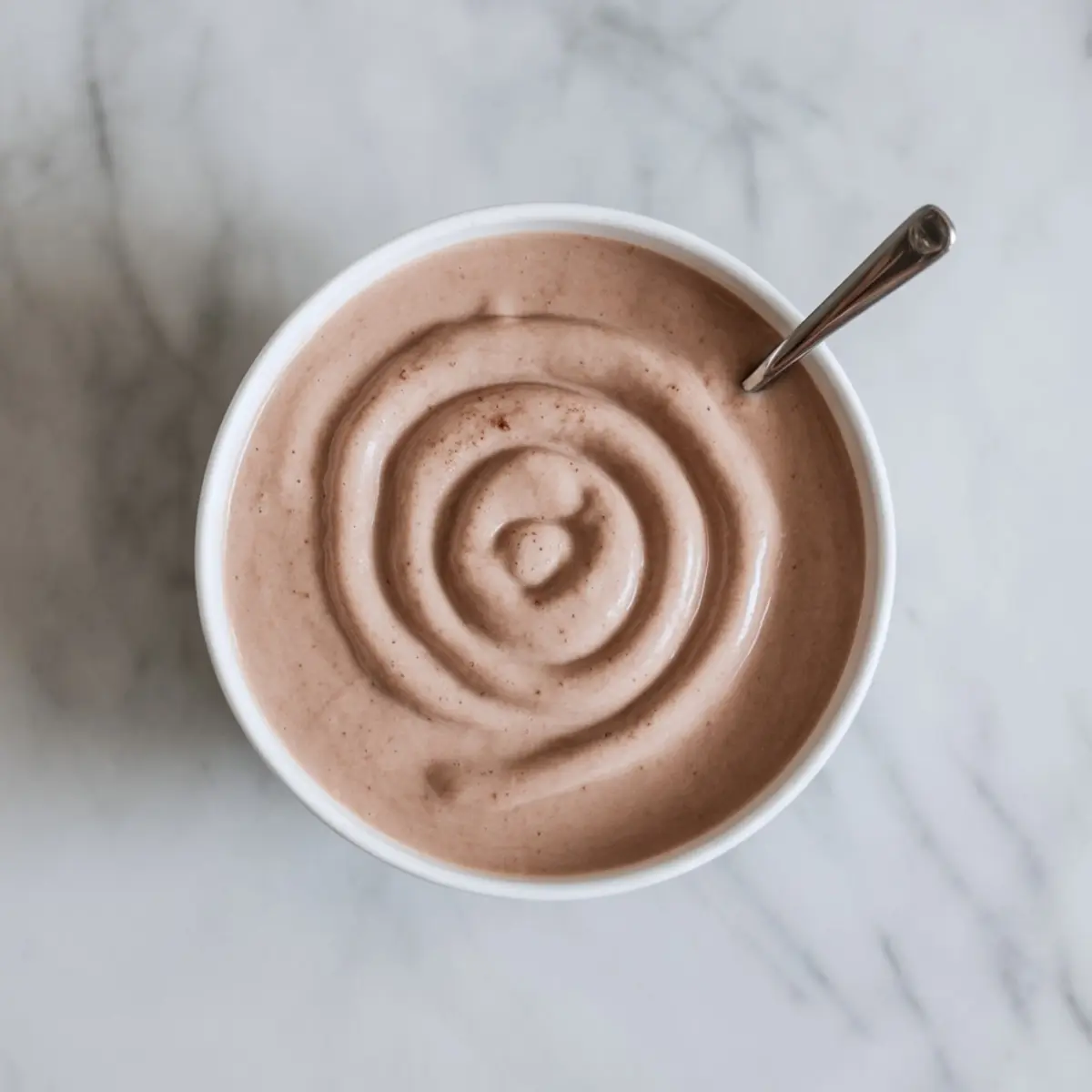 Top view of a creamy cinnamon banana smoothie blend with a smooth swirl pattern in a white bowl on a marble counter, featuring a metal spoon.