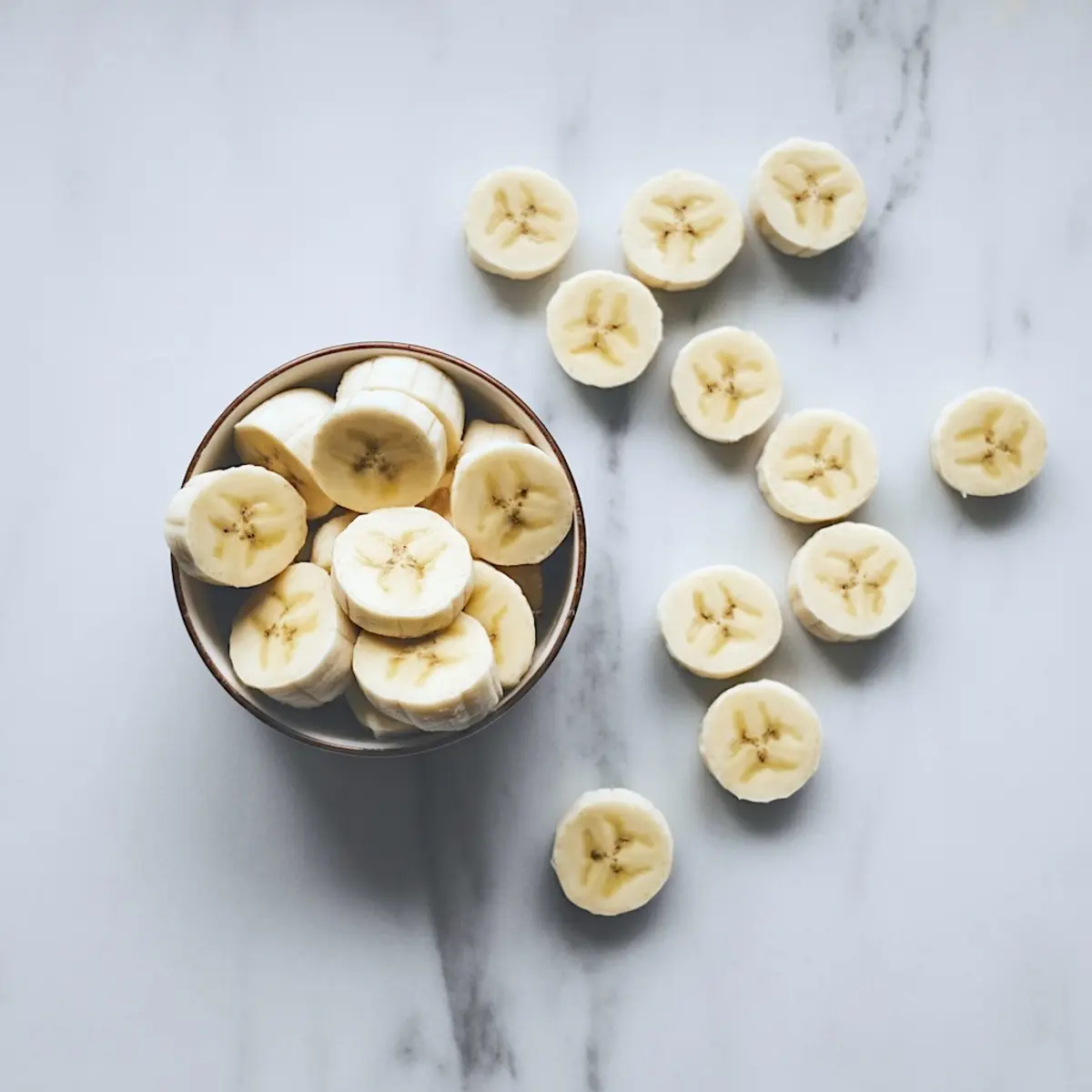 Overhead view of a bowl of sliced bananas with extra banana slices scattered on a white marble surface, highlighting the fruit’s soft texture and star-like core pattern.
