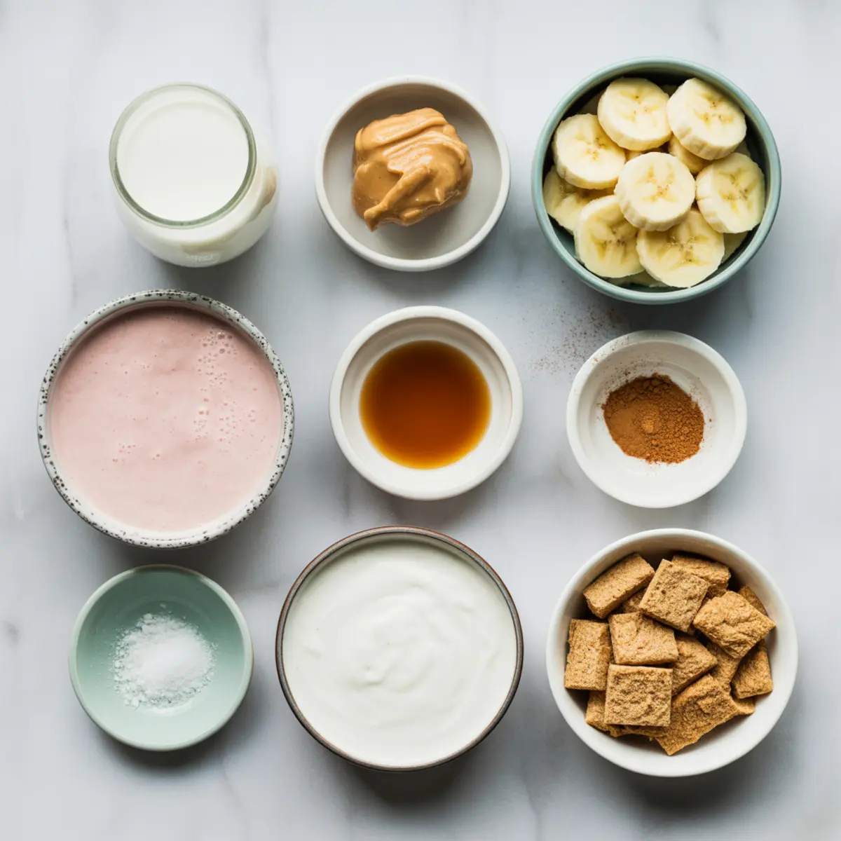 Flat lay of cinnamon roll smoothie bowl ingredients arranged in small bowls on a marble surface, including sliced banana, yogurt, peanut butter, vanilla extract, milk, cinnamon, cereal squares, salt, and pink smoothie base.