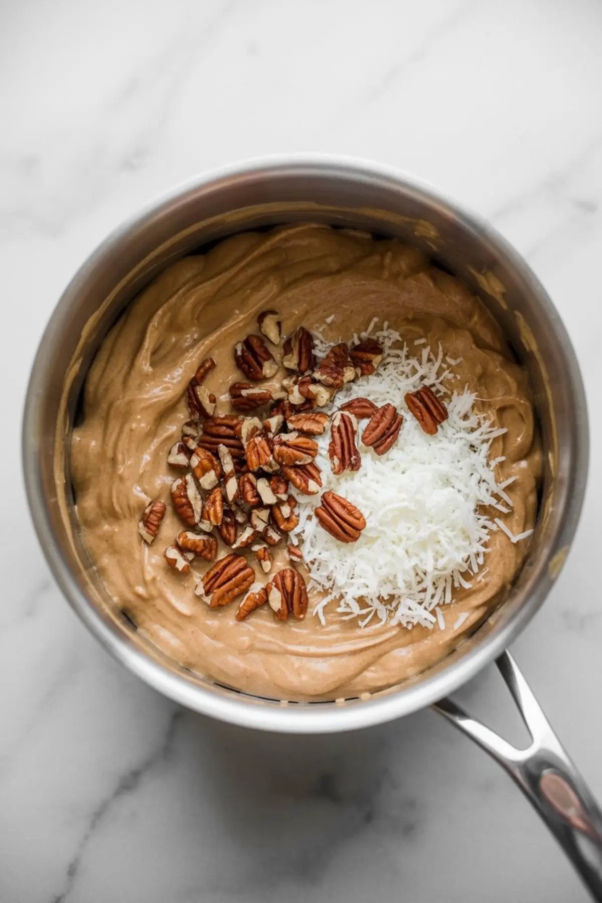 Coconut pecan frosting in a saucepan, topped with shredded coconut and chopped pecans ready to be mixed into the creamy base.