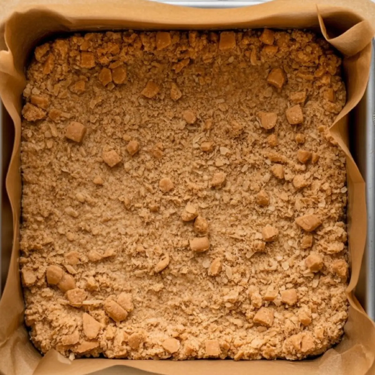 Square baking pan lined with parchment paper holding freshly baked coconut toffee bar base topped with a golden brown oat and toffee crumble.
