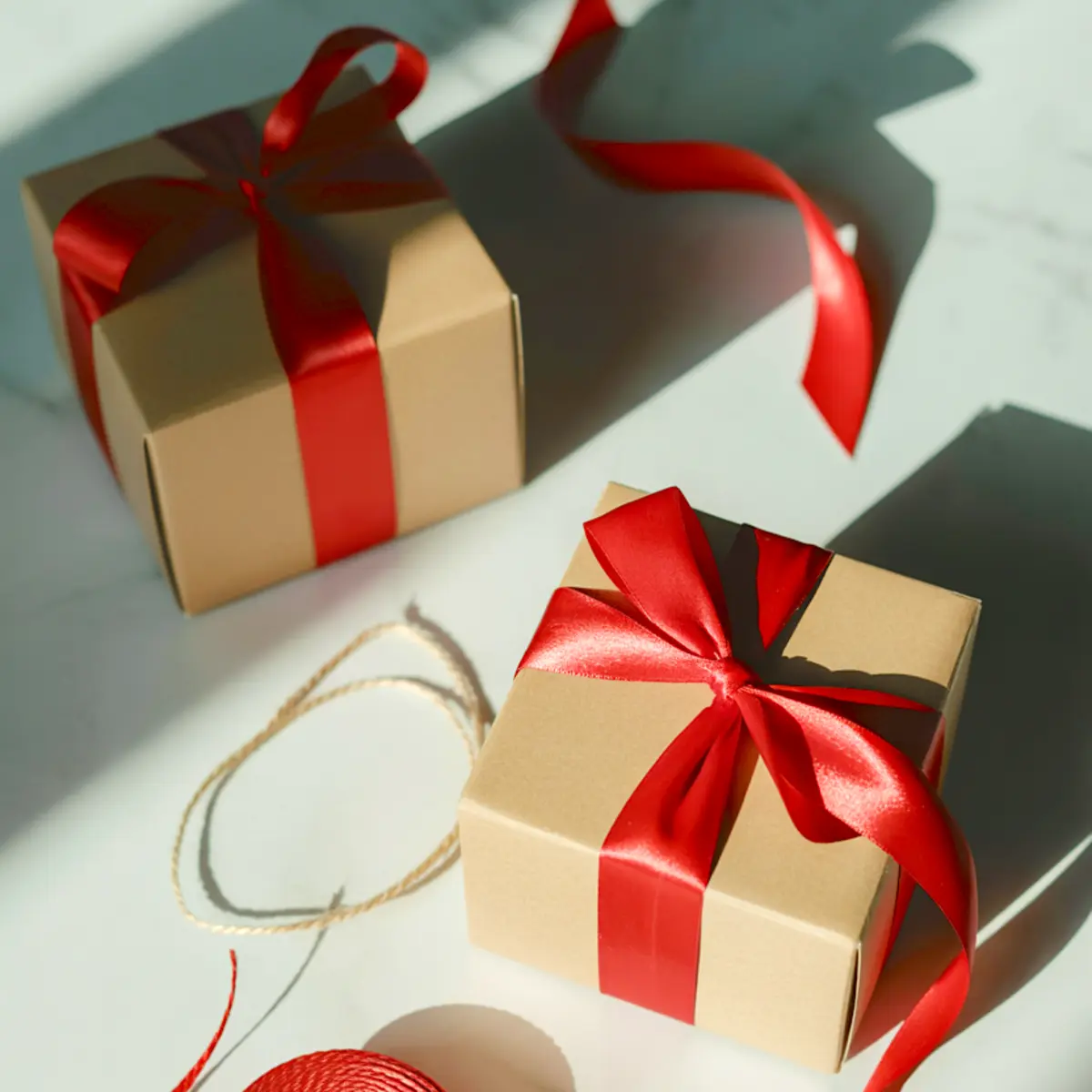 Two kraft gift boxes wrapped with shiny red satin ribbon bows, positioned on a white surface with soft sunlight and decorative twine nearby, ideal for Christmas cookie gift packaging or festive treat boxes.