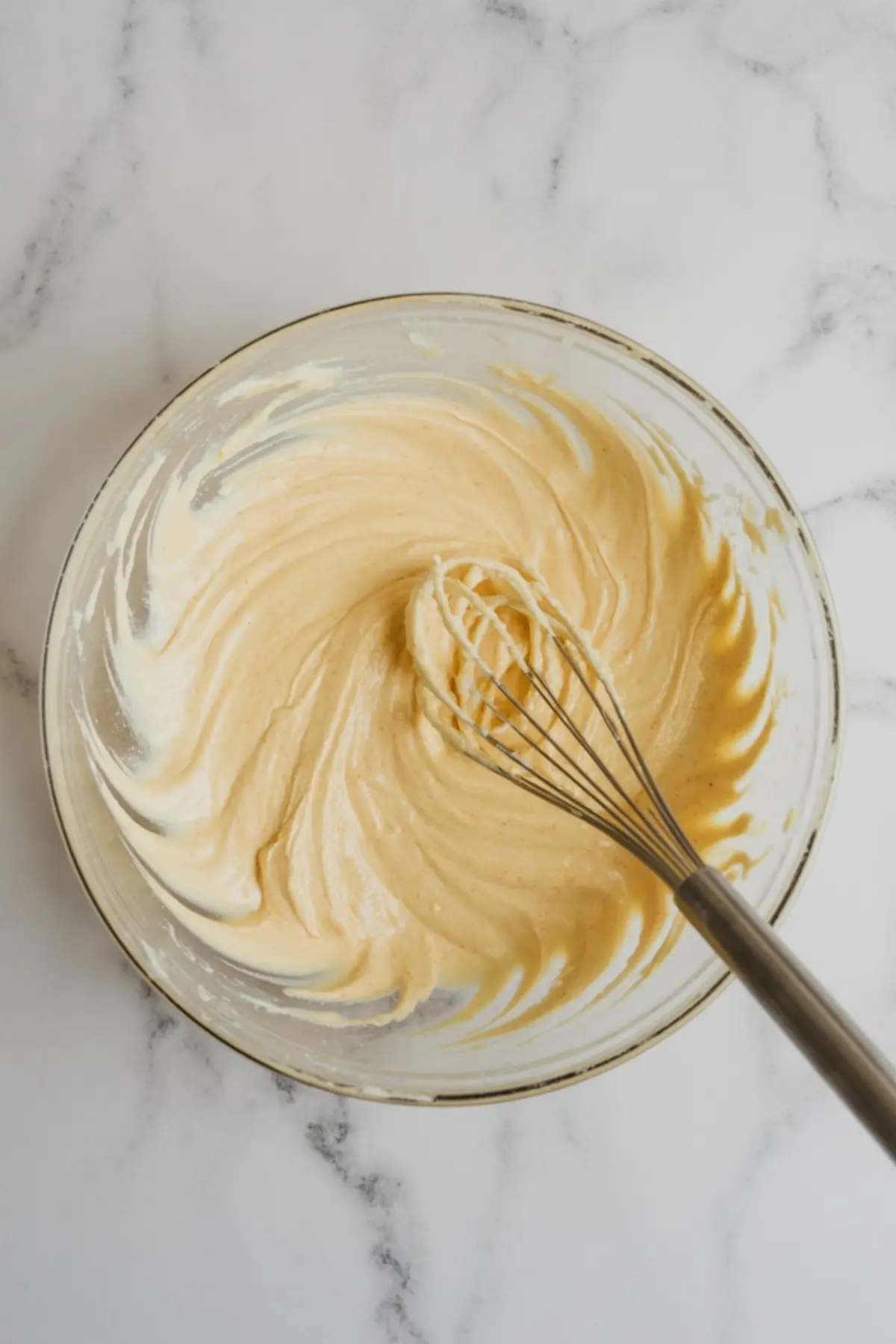 Smooth and creamy cheesecake filling being whisked in a glass bowl on a marble countertop, showing a rich texture ideal for tartlets or mini cheesecakes.