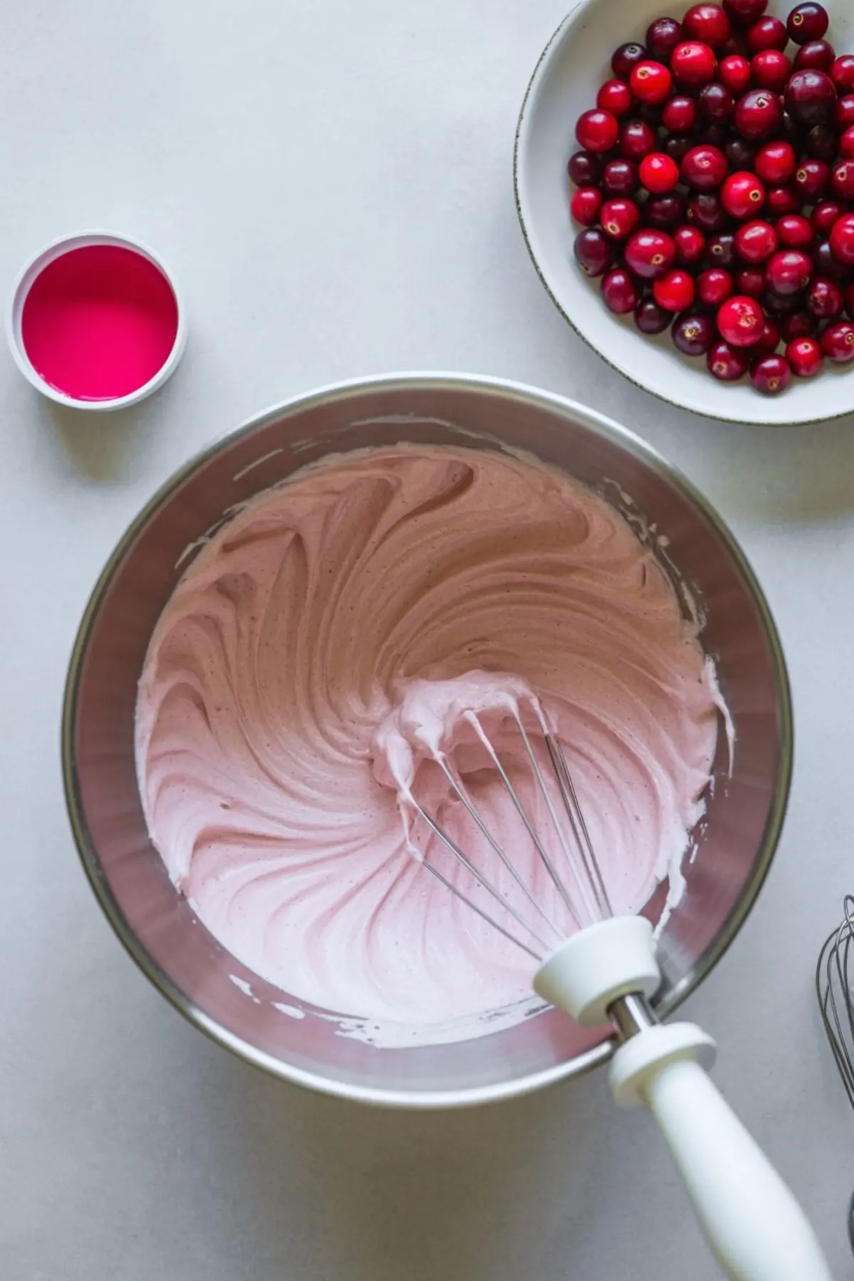 Stainless steel mixing bowl filled with fluffy pink cranberry mousse being whipped with a handheld whisk, surrounded by a bowl of fresh cranberries and a small container of vibrant cranberry juice on a light background.
