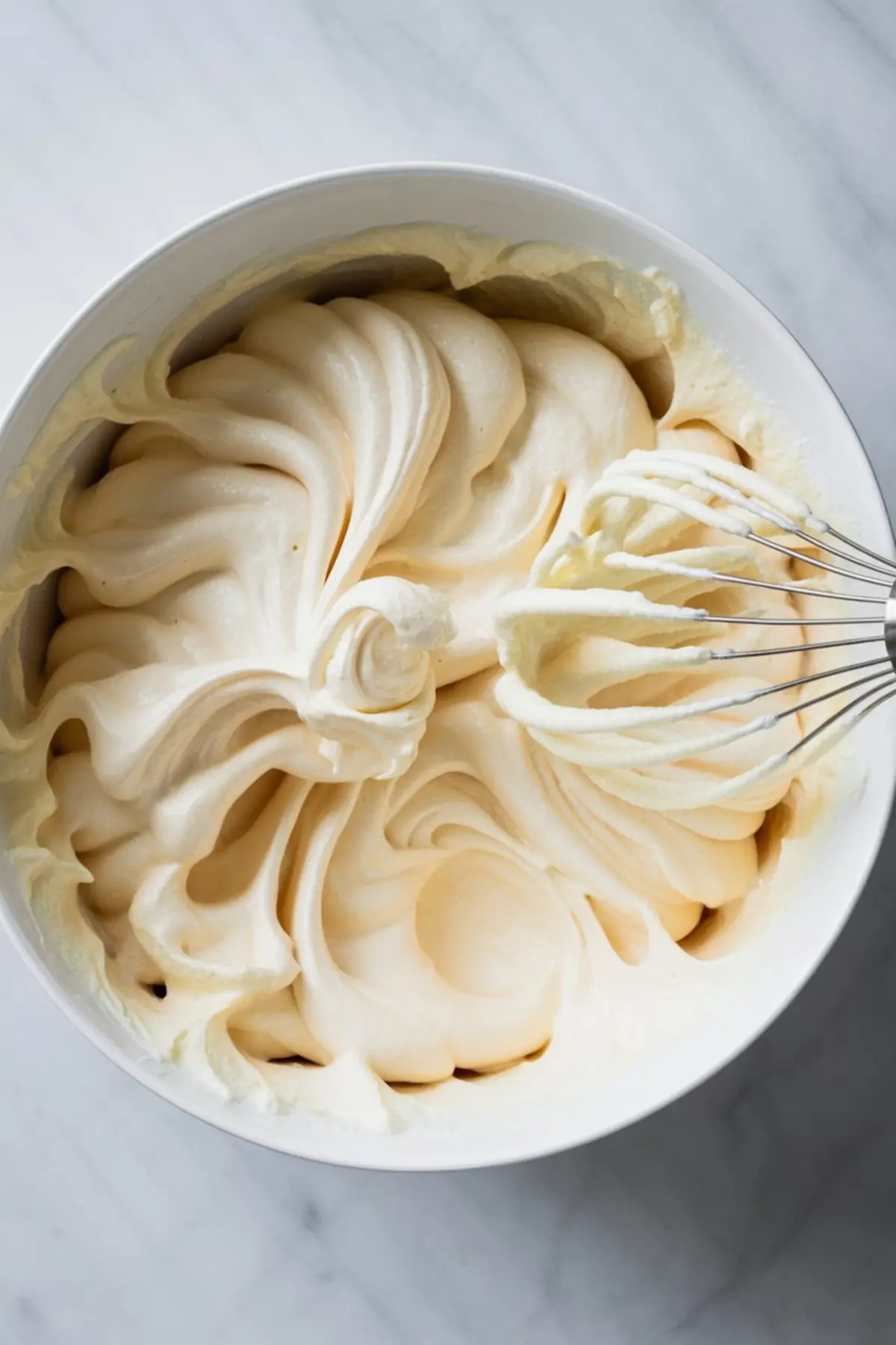 Creamy whipped vanilla mousse in a white mixing bowl with a whisk, showing thick, airy texture swirled into soft peaks on a marble countertop.
