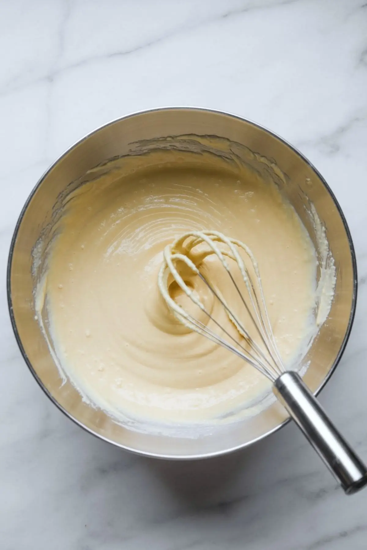 Metal mixing bowl filled with creamy cheesecake batter, with a stainless steel whisk resting inside on a white marble surface.

