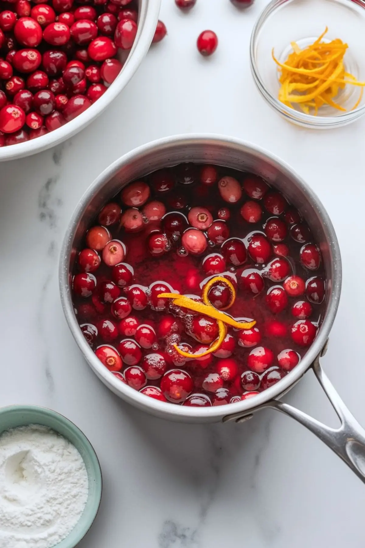 Saucepan filled with simmering cranberries and orange zest for cranberry sauce, surrounded by fresh cranberries, powdered sugar, and a small bowl of orange zest.
