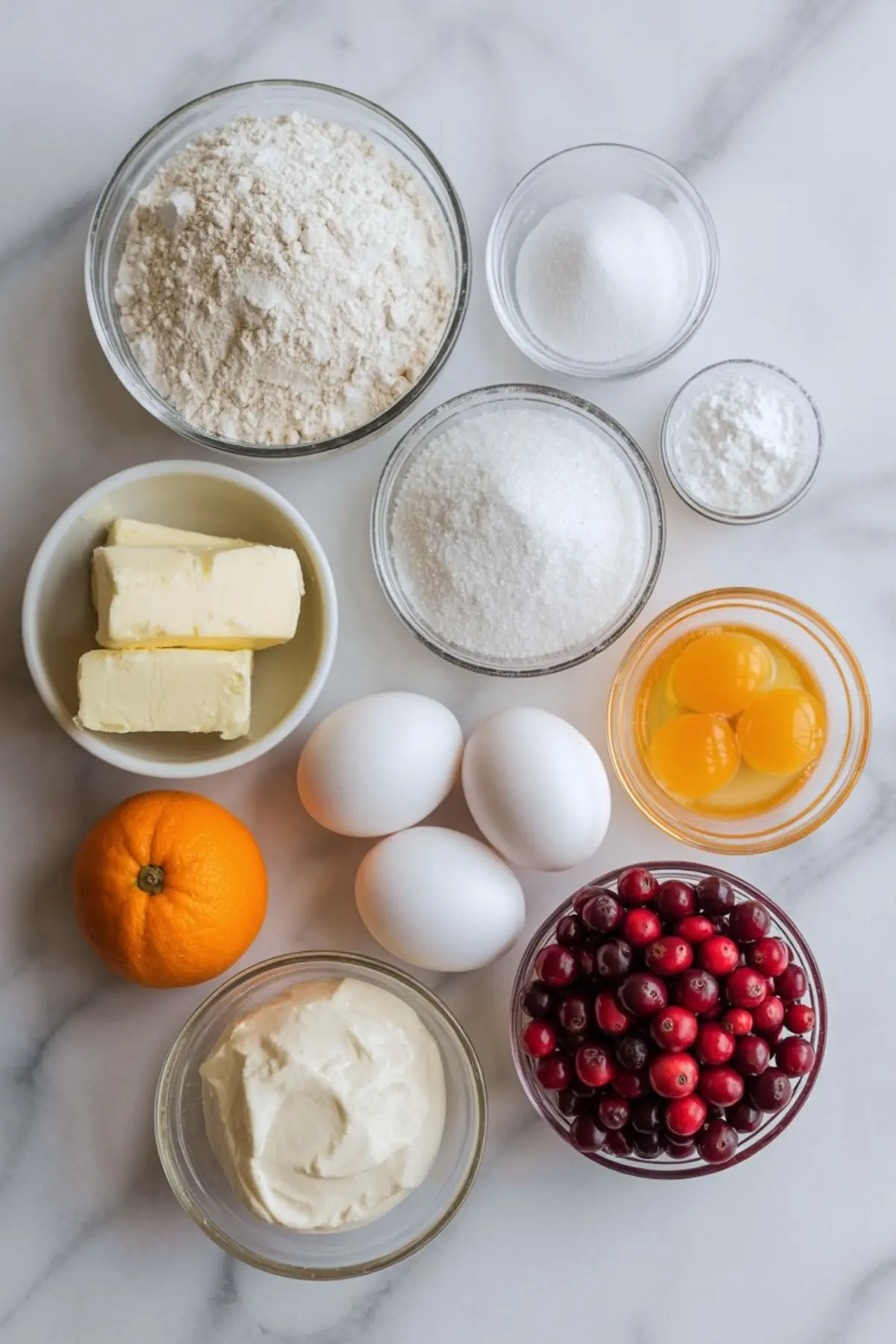 Flat lay of baking ingredients in glass bowls, featuring flour, sugar, butter, eggs, orange, cranberries, and sour cream arranged on a white marble surface.
