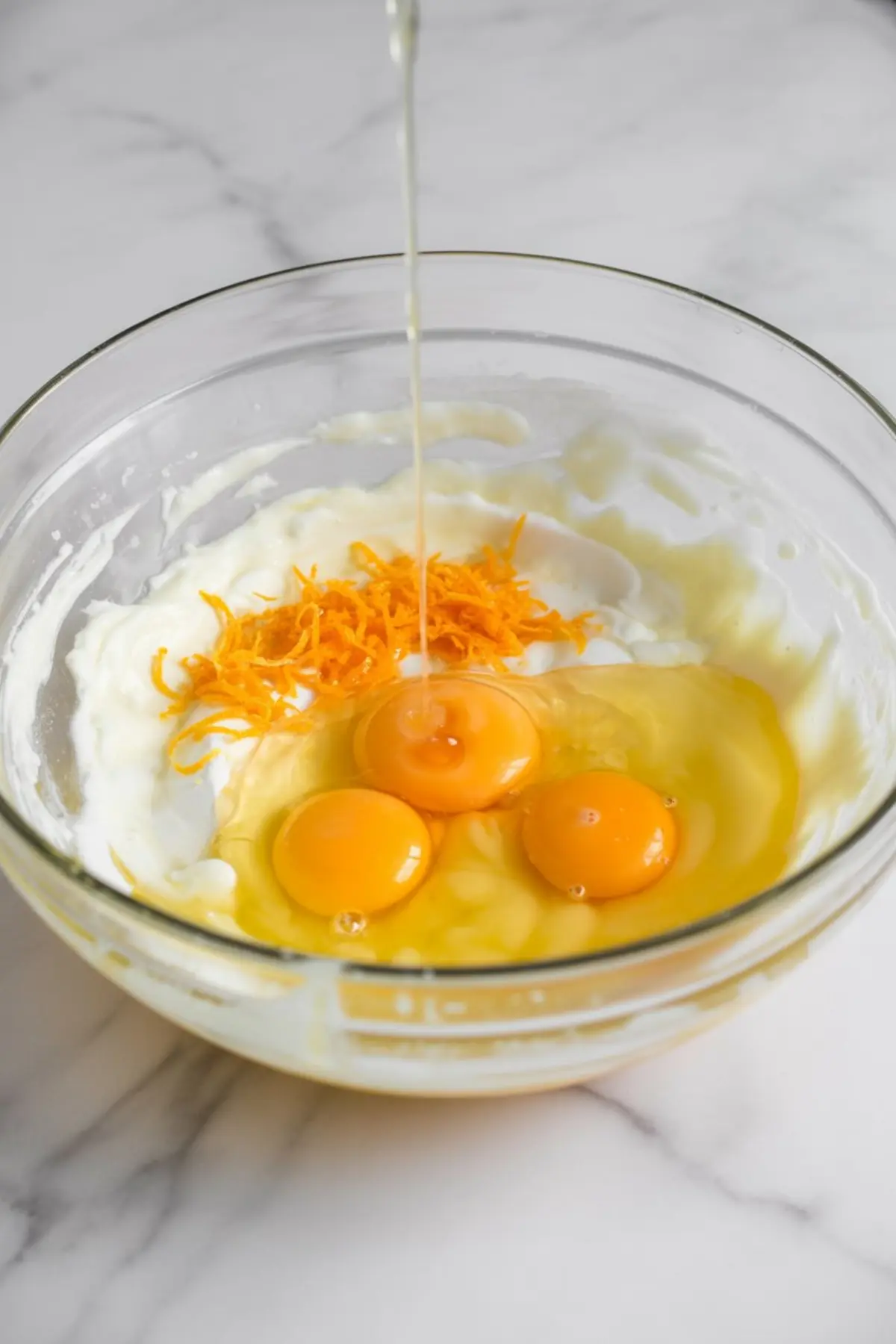 Glass mixing bowl containing eggs, orange zest, and a creamy wet ingredient mixture with a stream of oil being poured in, set on a white marble countertop.
