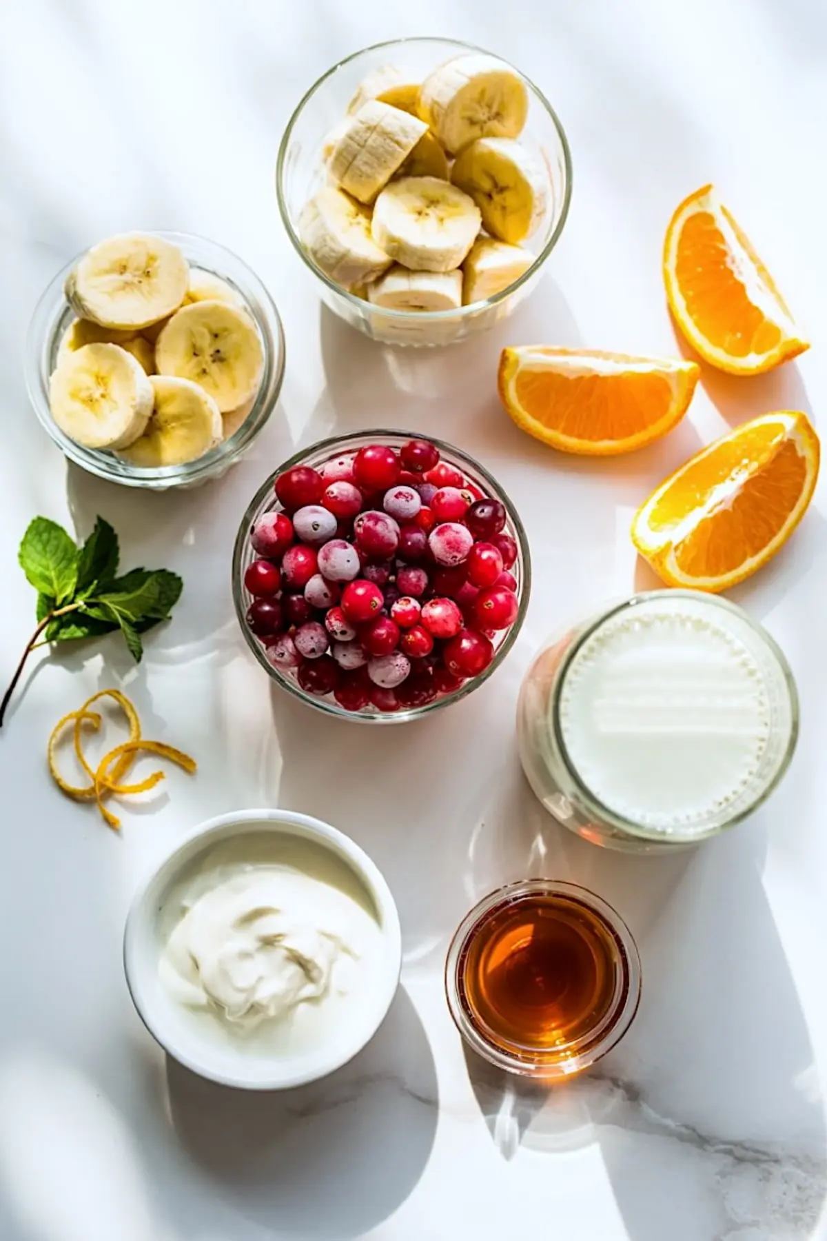Flat lay of fresh smoothie ingredients including banana slices, orange wedges, frozen cranberries, Greek yogurt, maple syrup, milk, and fresh mint on a white surface under natural sunlight.
