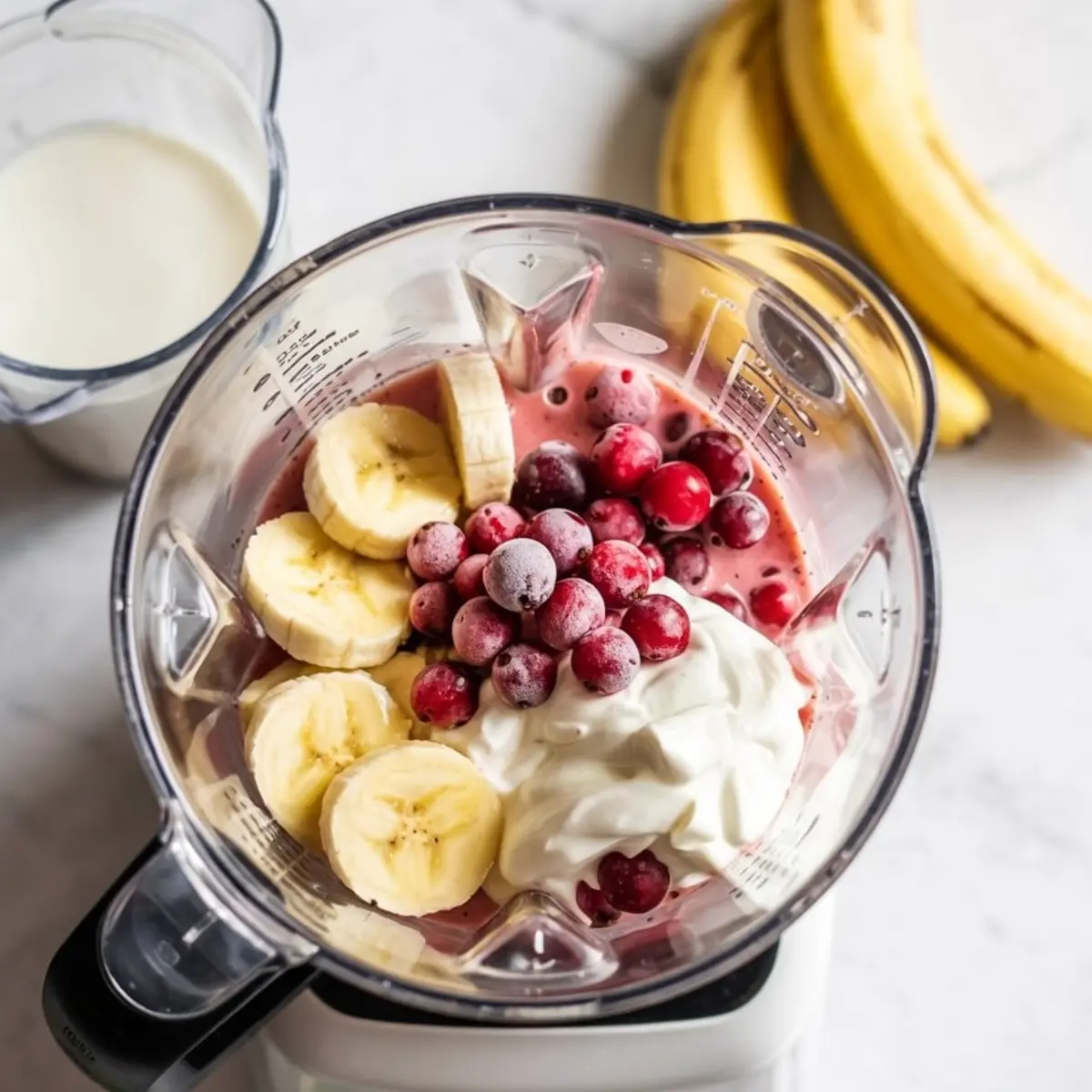 Top view of a blender filled with banana slices, frozen cranberries, Greek yogurt, and a pink smoothie base, with bananas and a cup of milk in the background, highlighting wholesome smoothie ingredients.
