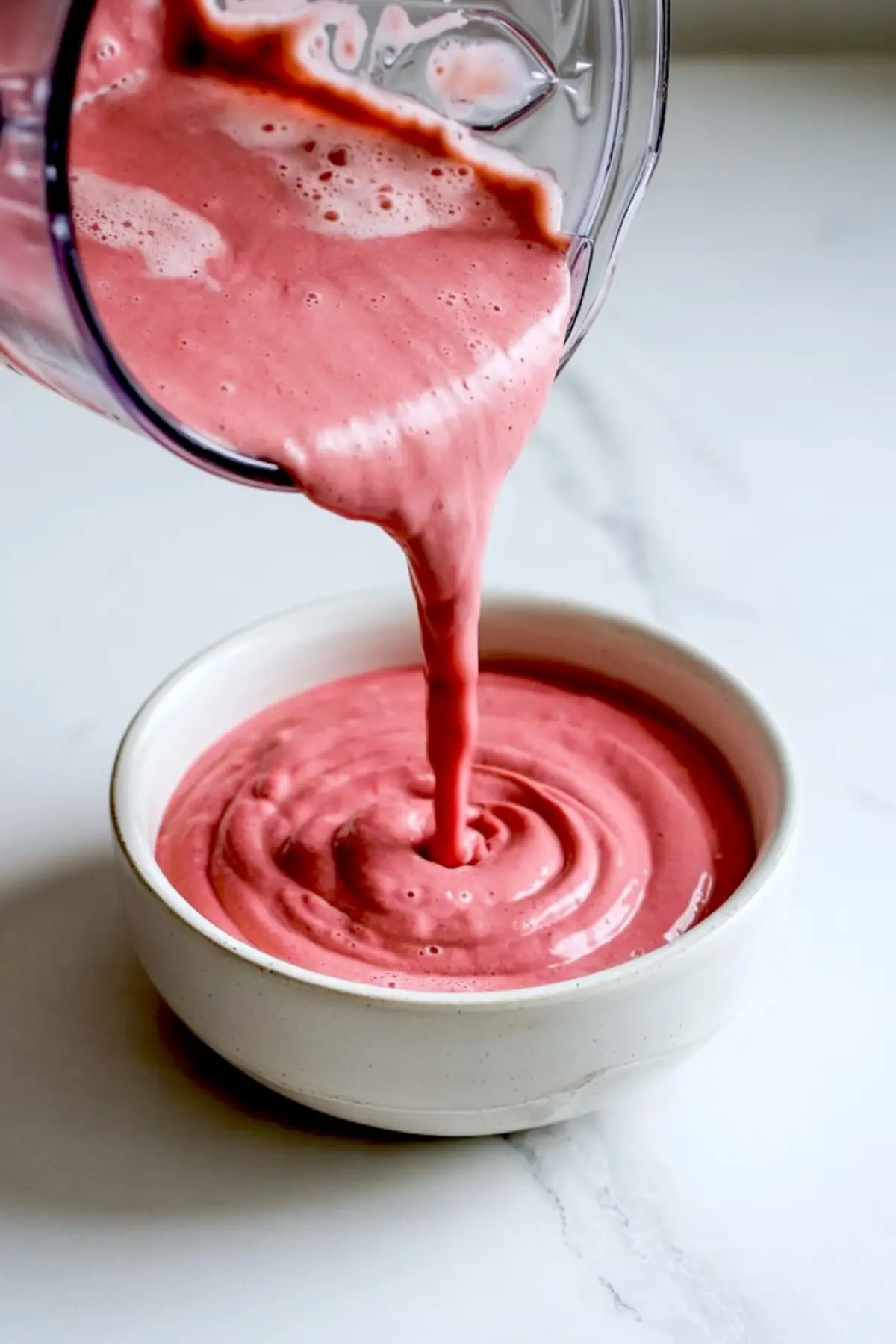 Thick pink smoothie being poured from a blender into a white ceramic bowl on a marble surface, showcasing a creamy texture ideal for smoothie bowl preparation or frozen fruit recipes.
