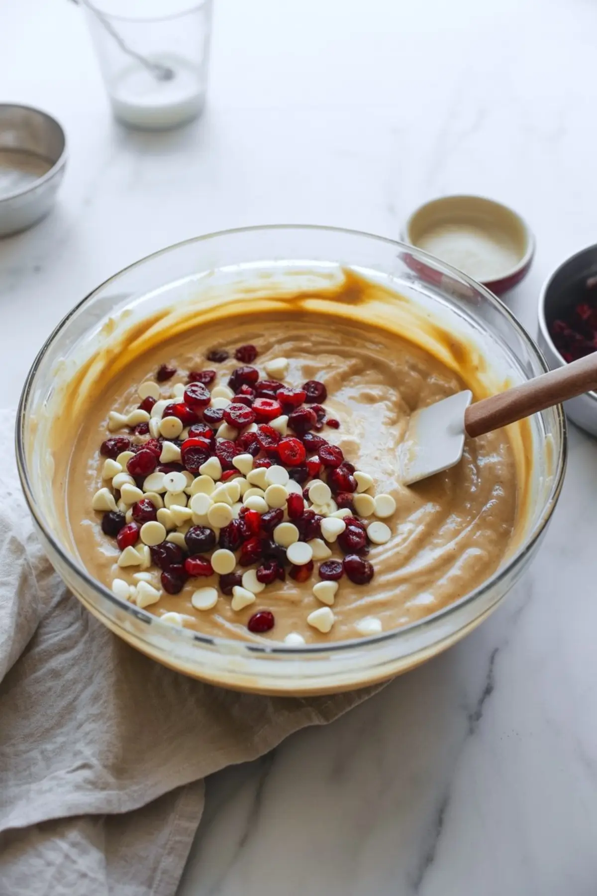 Glass mixing bowl filled with smooth batter topped with white chocolate chips and dried cranberries, with a rubber spatula resting inside.