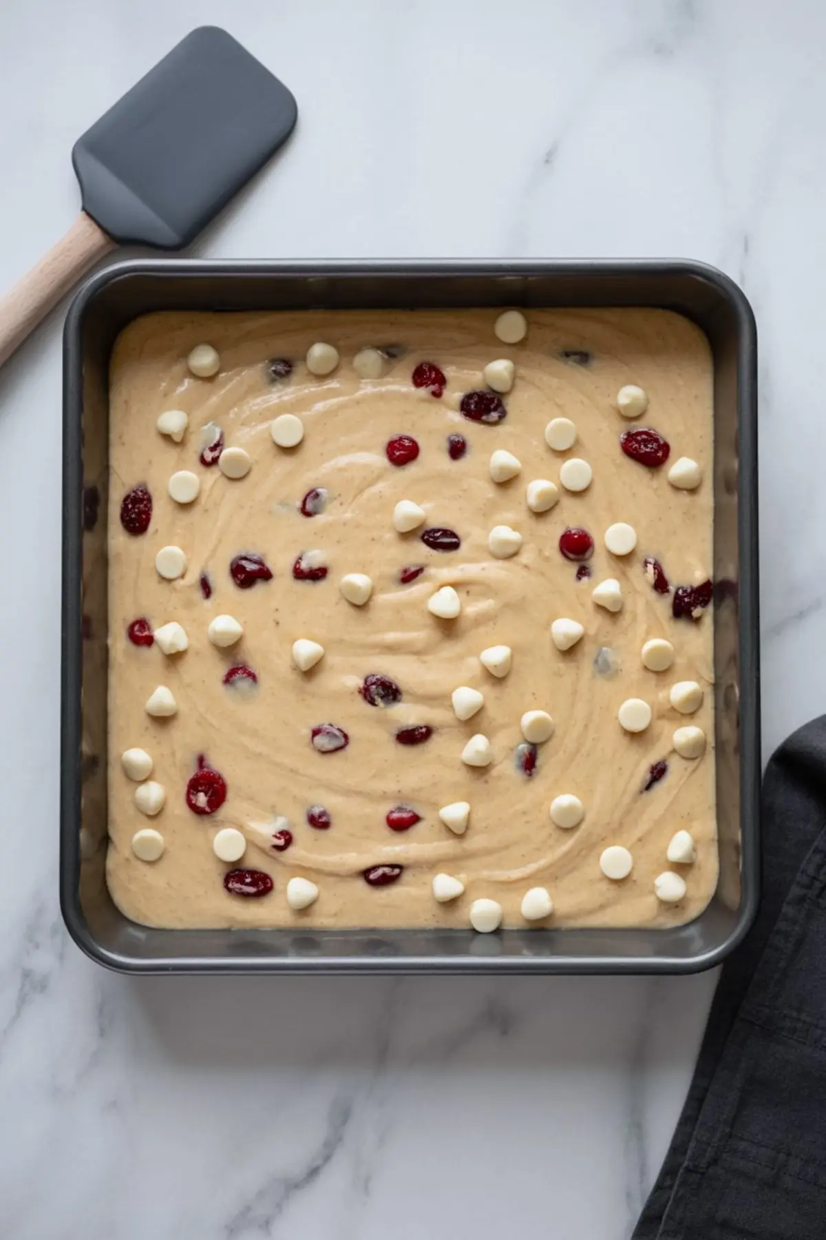 Square baking pan filled with unbaked cranberry white chocolate blondie batter, topped with scattered white chocolate chips and cranberries.