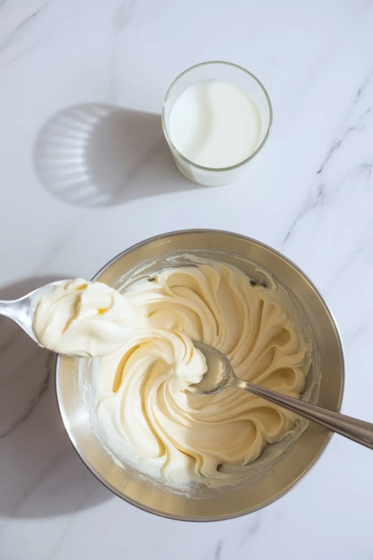 Spoonful of fluffy cream cheese frosting being held above a metal bowl, with a glass of milk in the background on white marble.
