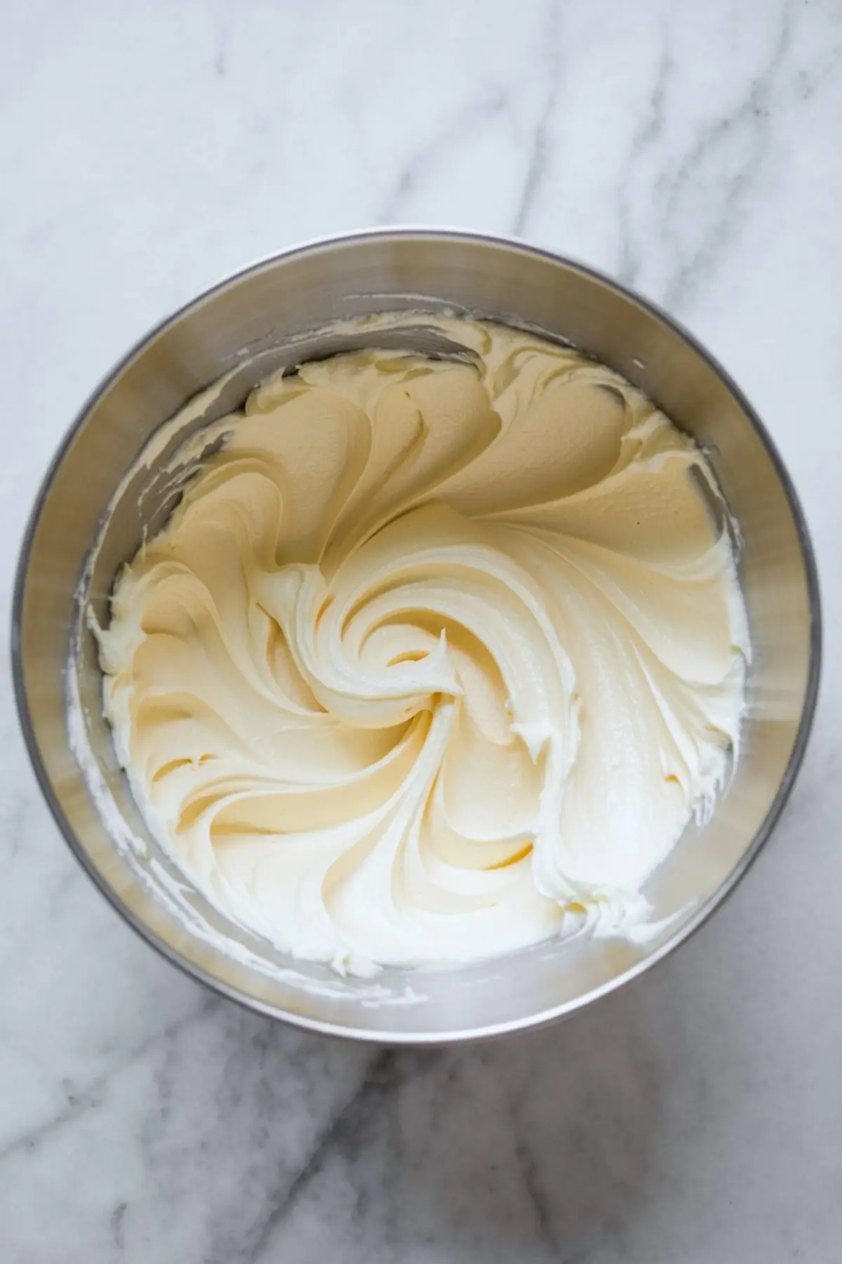 Whipped cream cheese frosting in a mixing bowl with smooth, swirled texture and soft peaks on a marble surface.
