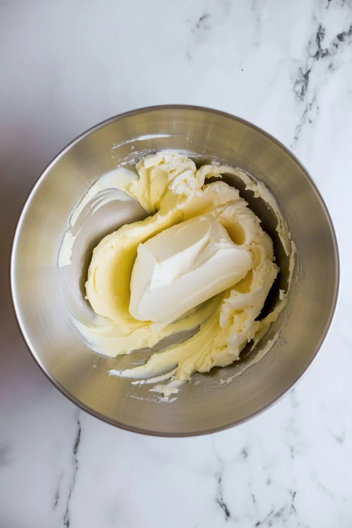Partially mixed butter and cream cheese in a metal bowl, showing the early stage of blending for cream cheese frosting on a marble background.
