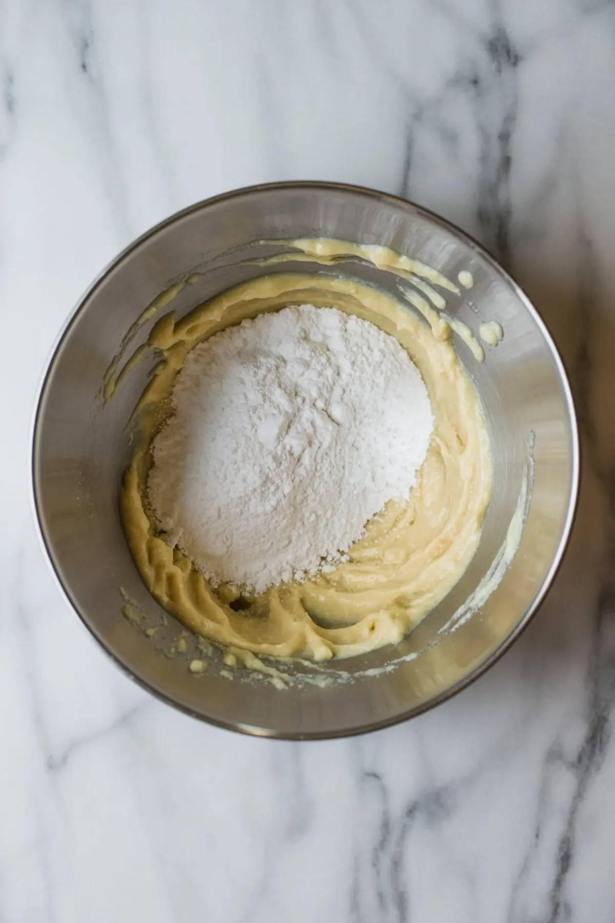 Stainless steel mixing bowl filled with thick yellow batter, topped with a heap of powdered sugar, placed on a white marble countertop.

